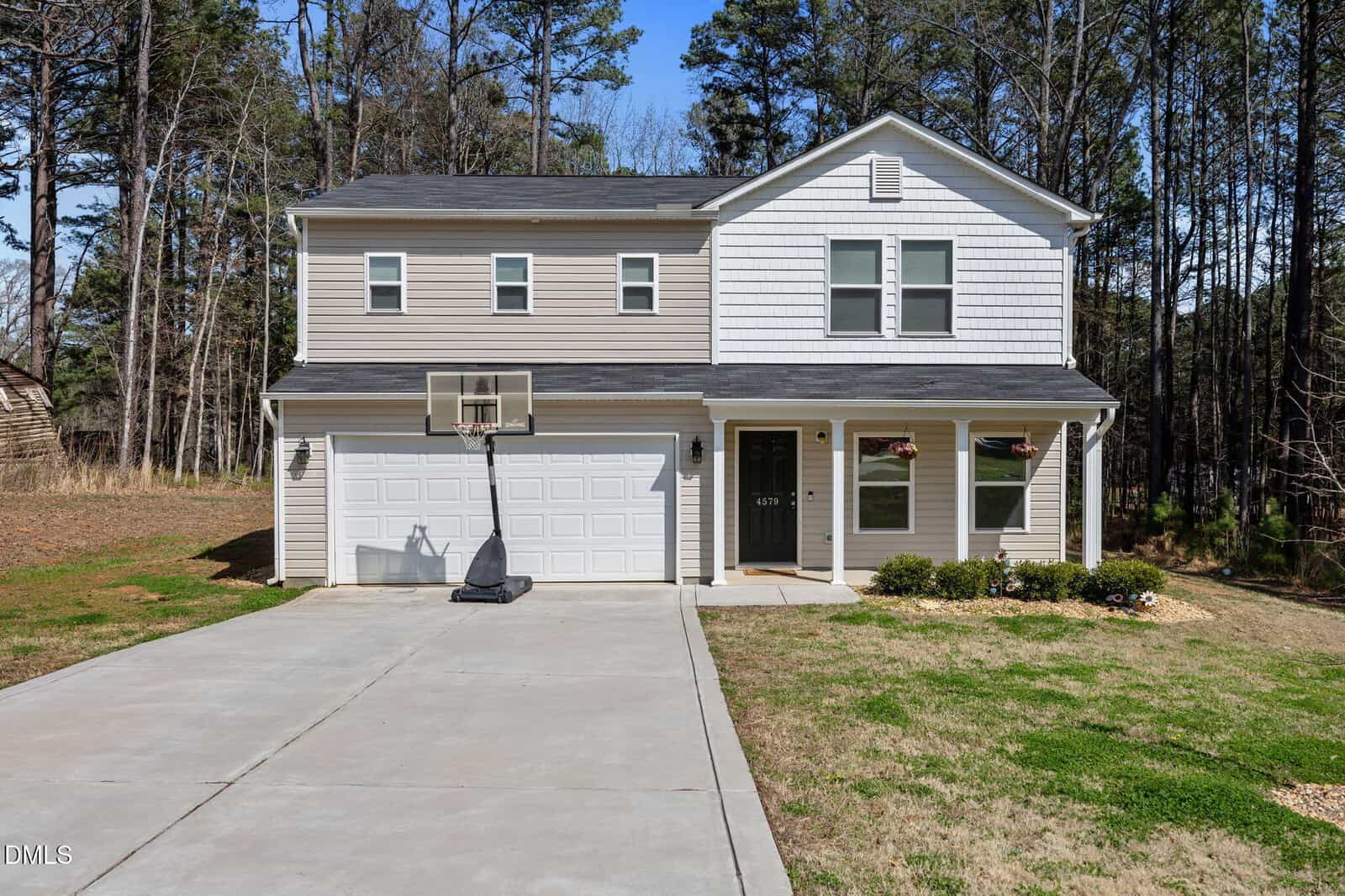 Two-story beige house with a double garage, basketball hoop, and a concrete driveway in the desirable 4579 Ashley View neighborhood of Oxford. Small shrubs adorn the front yard, while tall trees provide a scenic backdrop.