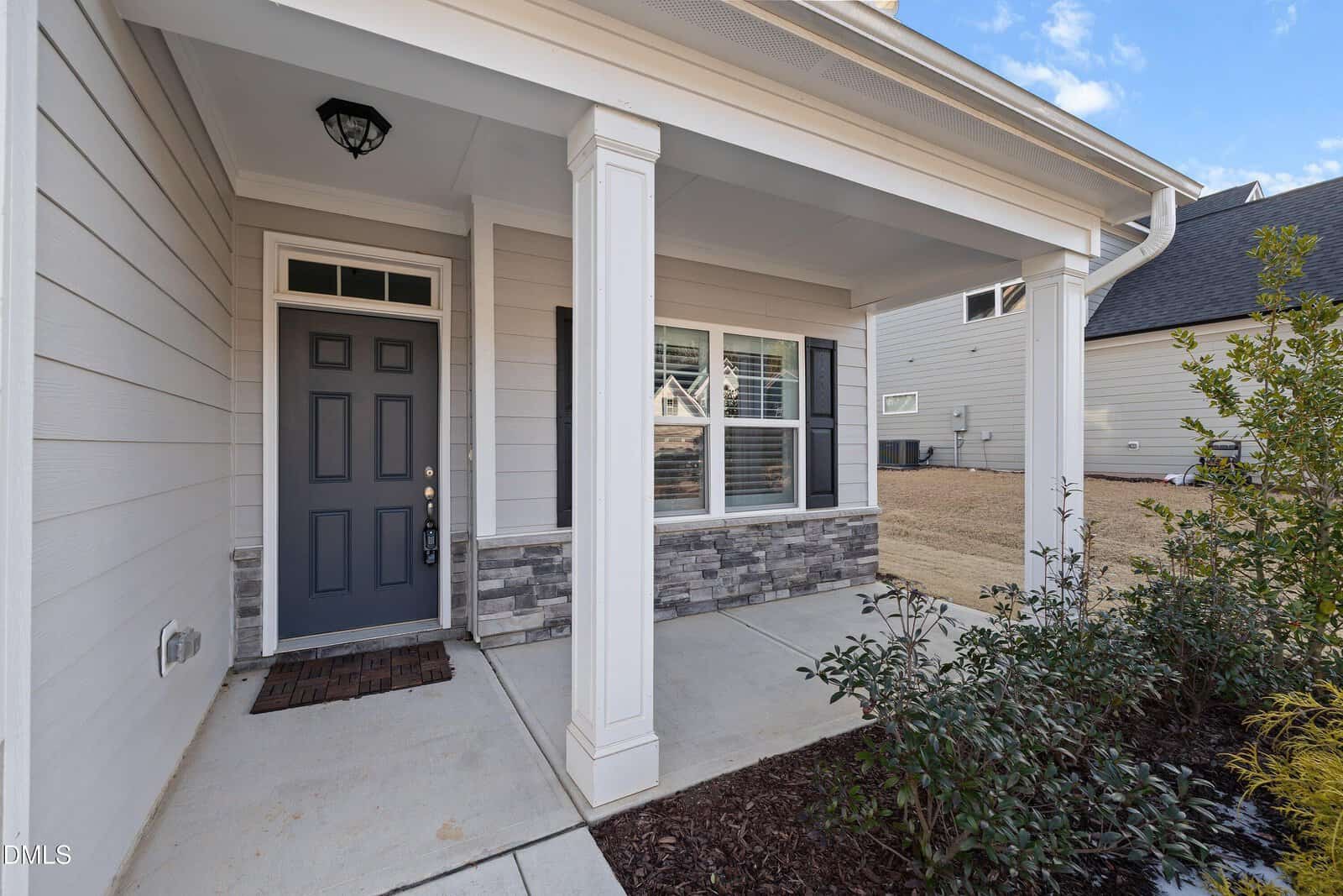 Front porch of a house with gray siding, a black door, white trim, two white columns, a welcome mat, stone detail below the window, and a landscaped area with shrubs; adjacent houses are visible.