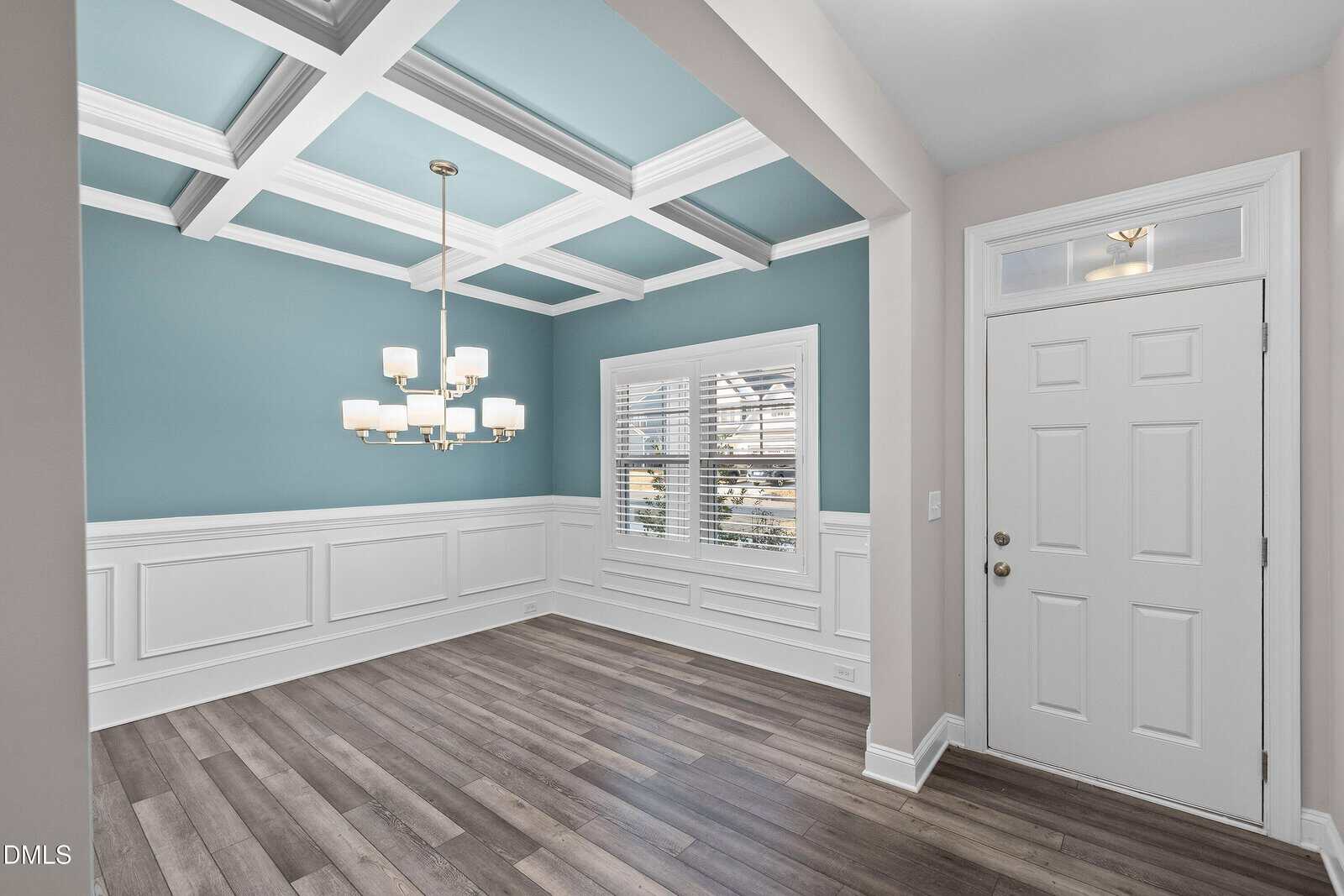 A bright entryway with a white door and a window, leading to a dining area with light blue walls, white wainscoting, a coffered ceiling, chandelier, and wood-look flooring. Sunlight comes through shuttered windows.