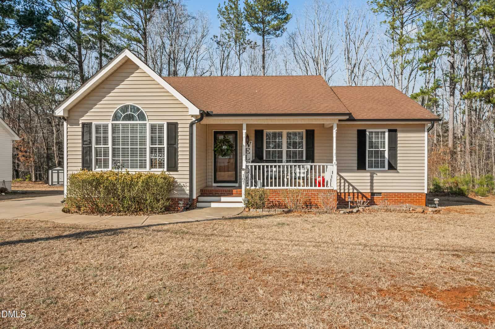 Single-story beige house with black shutters, a brown shingled roof, front porch with white railing, and a small concrete walkway. The lawn is dry and surrounded by trees with no leaves.