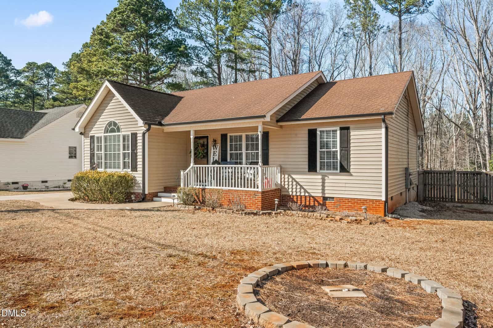 Single-story beige house with a gable roof, white trim, and small covered front porch. The yard is mostly dry grass with a circular stone-bordered area in front. Trees and neighboring homes are visible in the background.