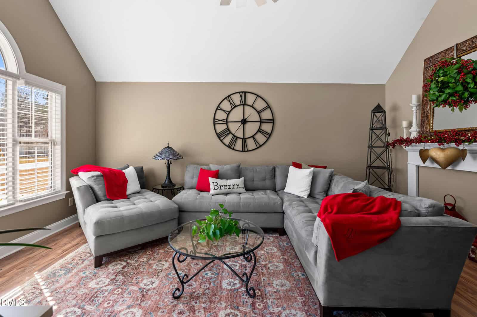 A living room with a large gray sectional sofa, red and white pillows and throws, a round glass coffee table with a plant, a patterned rug, a wall clock, and a decorated white mantel against a tan wall.