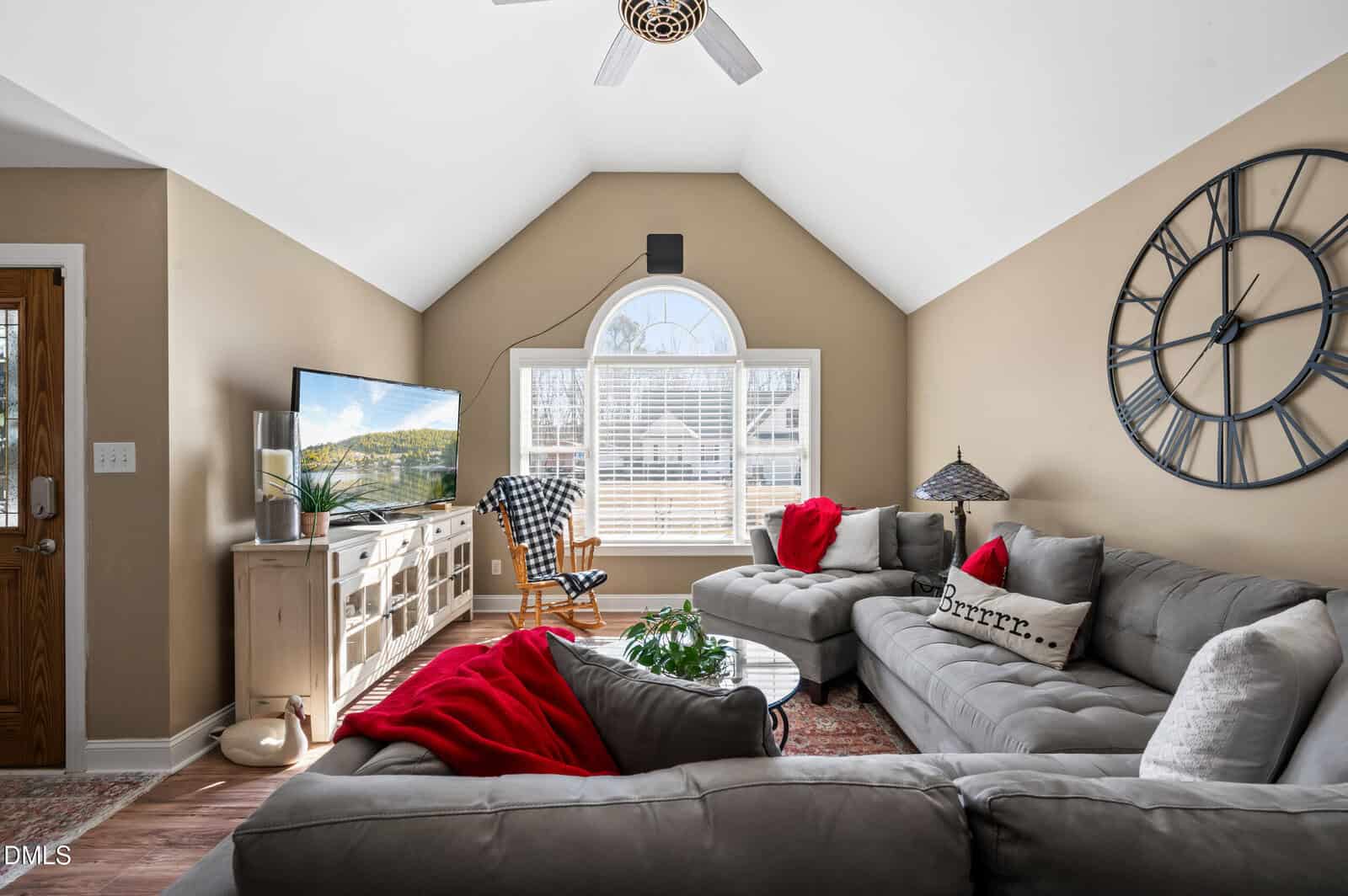A living room with vaulted ceiling, beige walls, two gray sectional sofas with red accents, a round coffee table, a large wall clock, a TV on a light wooden stand, and a window letting in natural light.