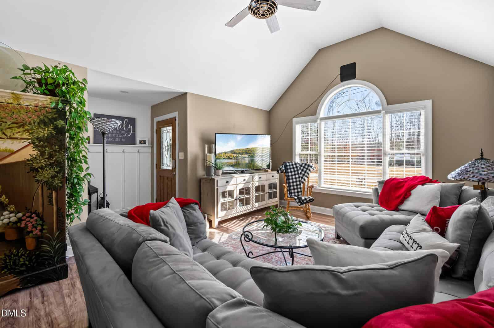 A bright living room with gray couches, red accents, and a TV on a white console. Large arched windows let in natural light. A round coffee table and decorative plants are also visible.