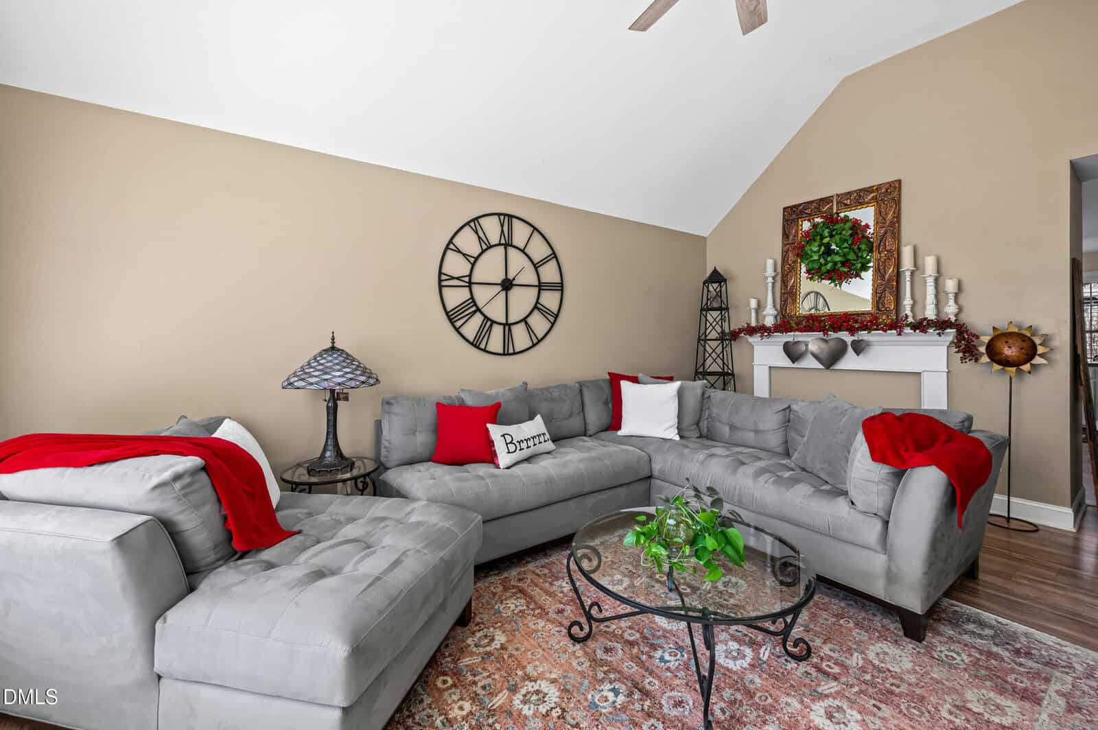 Living room with gray sectional sofas, red and white pillows, red blankets, a round glass coffee table, a decorative wall clock, and a white mantel with a mirror, wreath, and ornaments. Rug covers wood floor.
