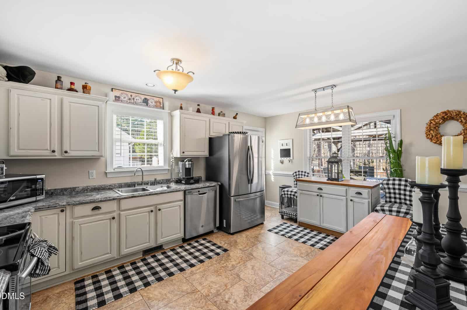 Bright kitchen with light-colored cabinets, stainless steel appliances, a center island, checkered rugs, and checkered upholstered chairs by a window. Decorative items are on cabinets and a black candle holder is on a wooden table.