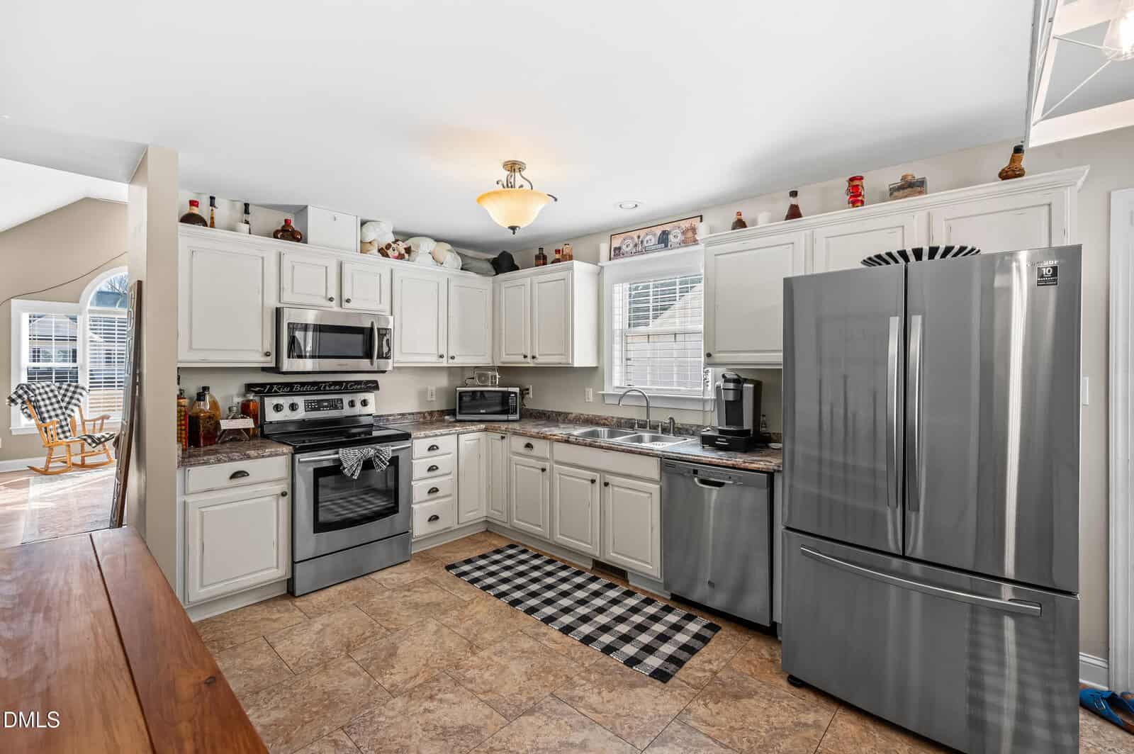 Modern kitchen with white cabinets, stainless steel appliances including a refrigerator, oven, microwave, and dishwasher. Granite countertops, a small checkered rug, and decorative items above the cabinets. Window brings in natural light.
