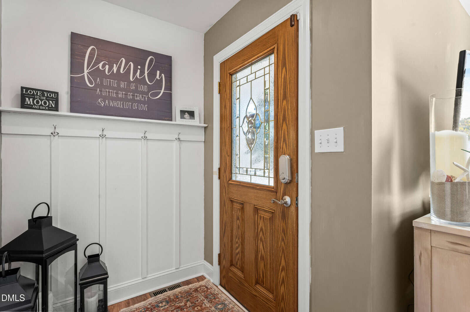A wooden front door with decorative glass is next to a beige wall with white trim. There are black lanterns on the floor, a "family" sign above white paneling, and a patterned rug on the floor.