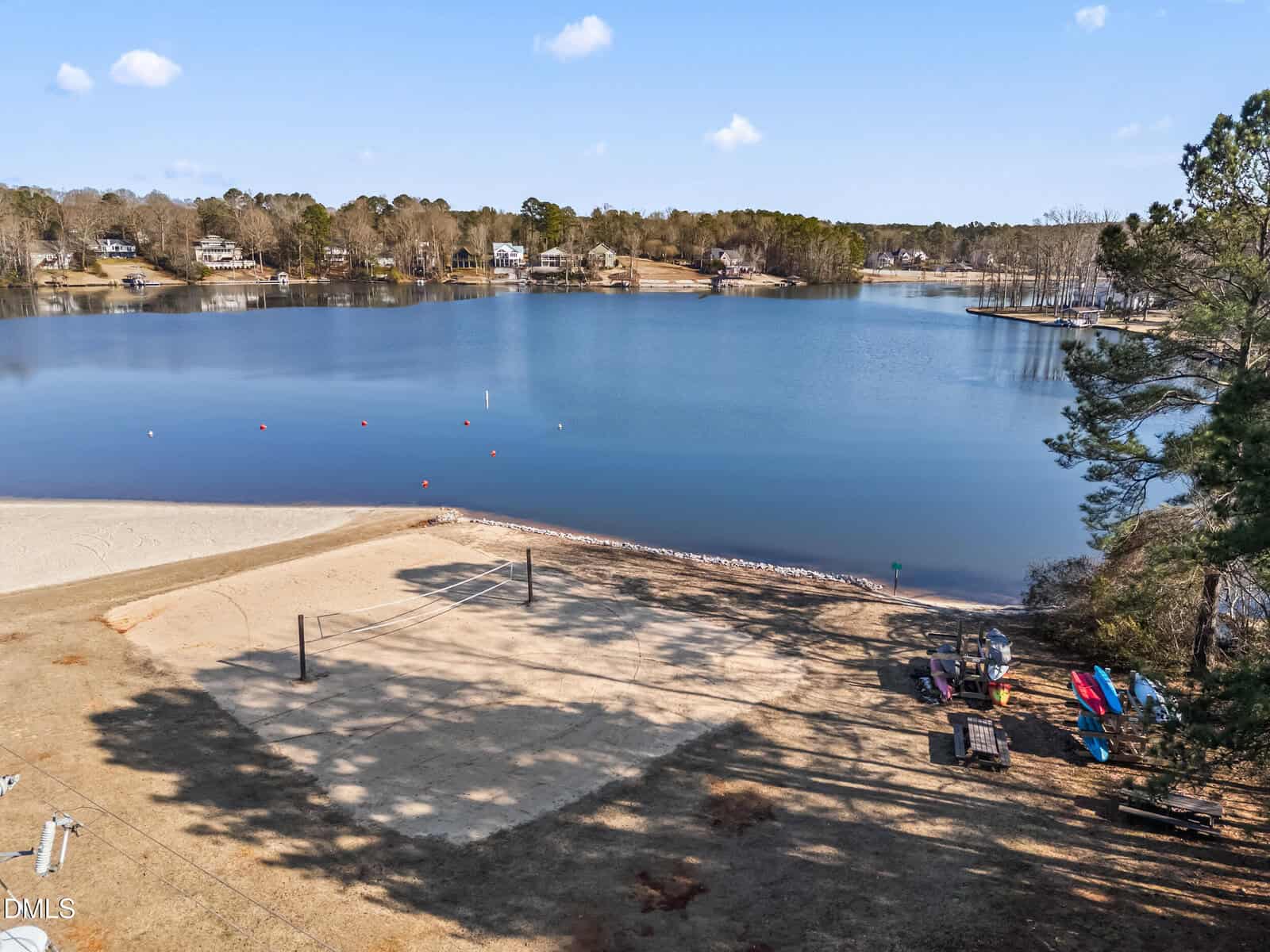 A sandy beach volleyball court is next to a calm lake surrounded by trees and houses. Several kayaks, canoes, and picnic tables sit on the shore under a clear blue sky.