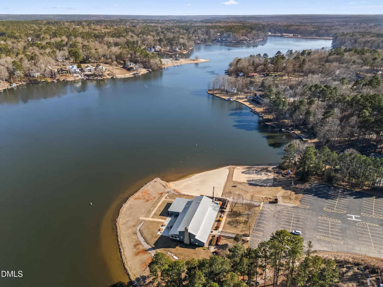 Aerial view of a lakeside area with a building near the shoreline, a sandy beach, a large empty parking lot, and surrounding wooded landscape. Houses are visible along the distant shoreline.