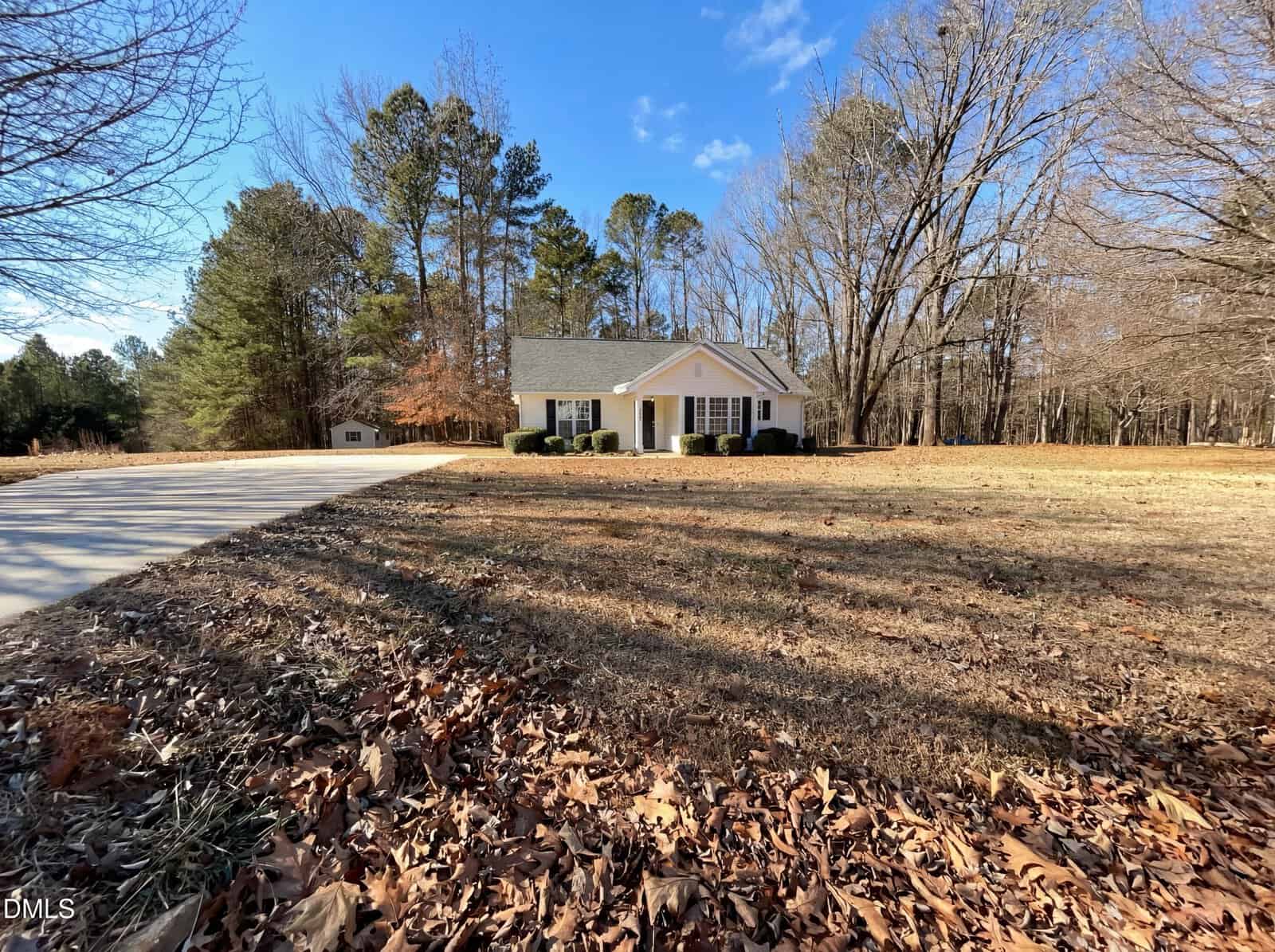 A single-story house with white siding and dark shutters sits on a large lot with a long driveway, surrounded by trees and dry grass under a clear blue sky.