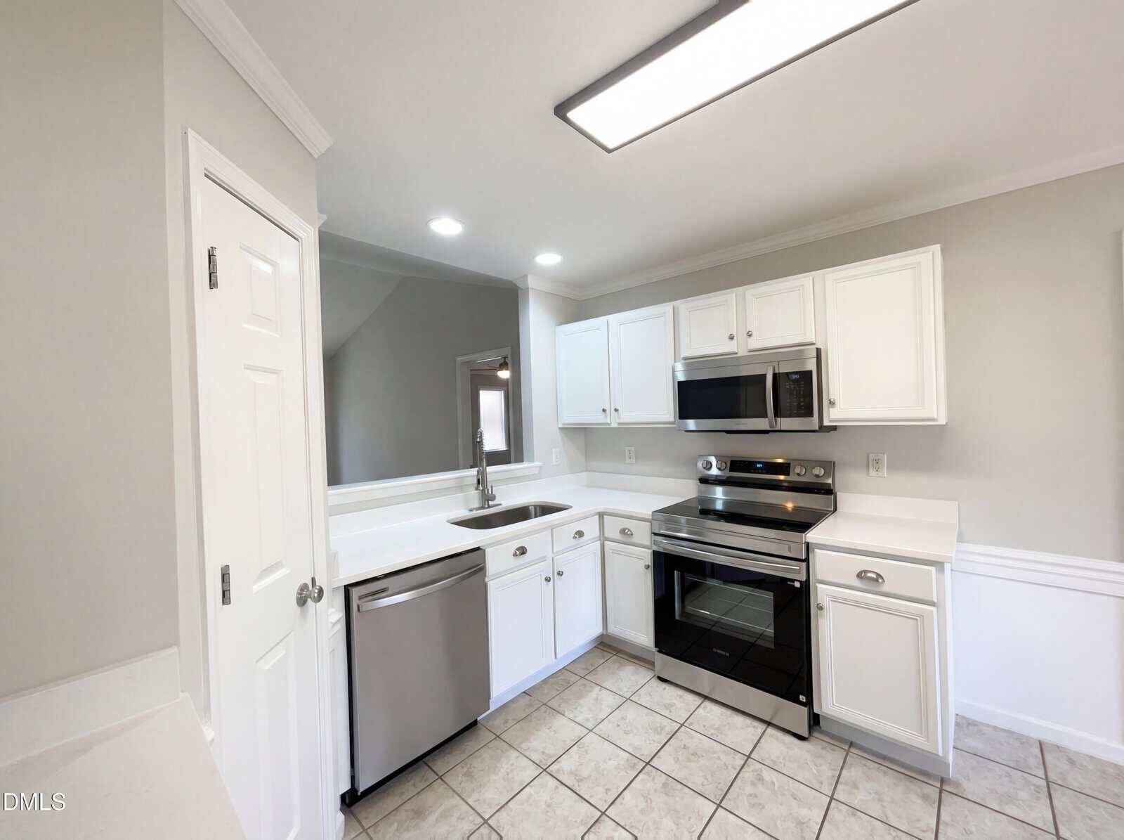 Modern kitchen with white cabinets, stainless steel appliances including a dishwasher, stove, and microwave, white countertops, double sink, tile floor, and neutral-colored walls. Fluorescent ceiling light overhead.