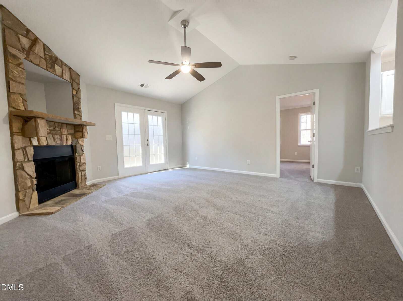 Spacious living room with light gray carpet, stone fireplace, ceiling fan, vaulted ceiling, and double glass doors leading outside. A doorway leads to another room, and natural light enters through multiple windows.