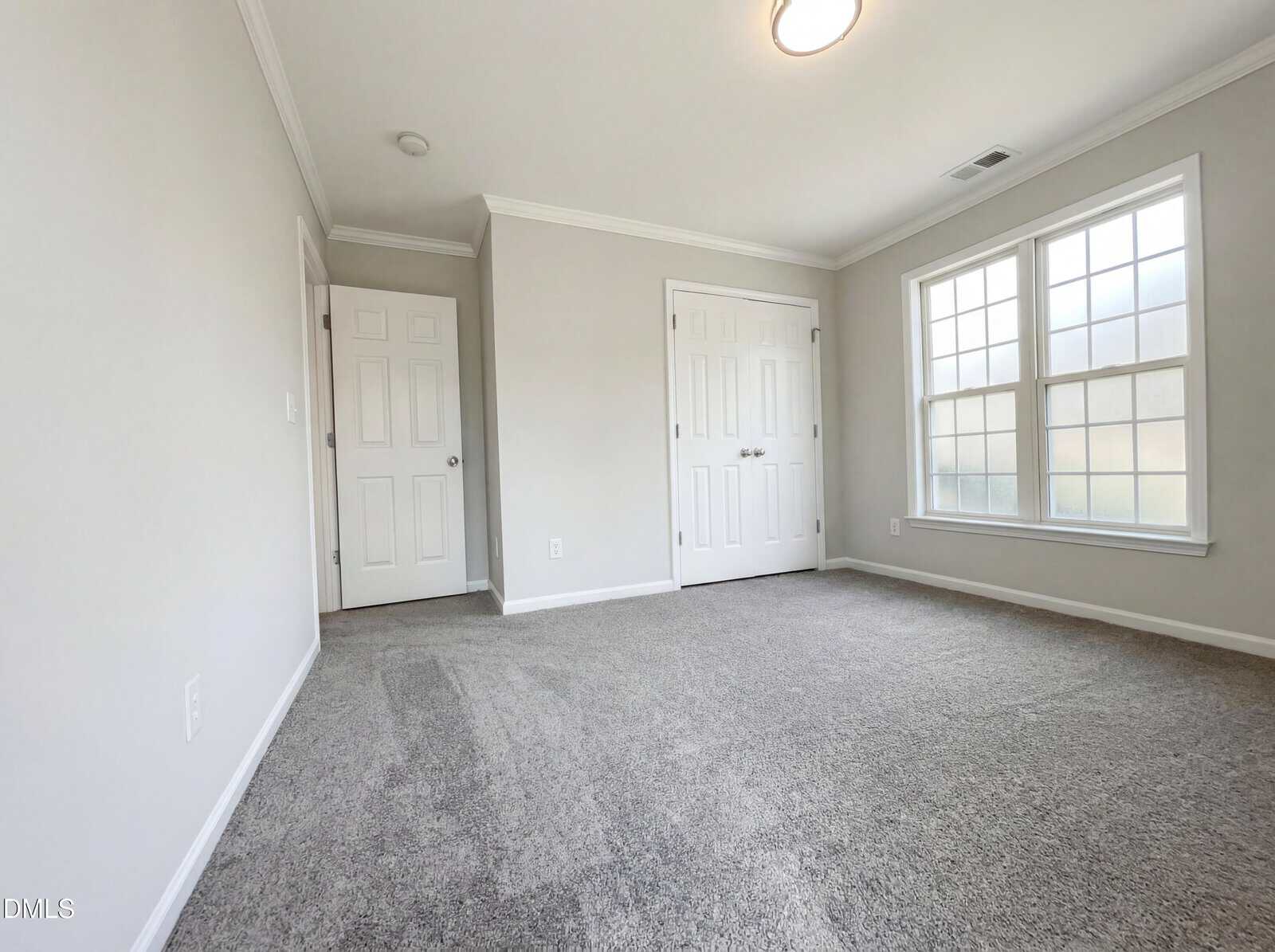 Bright, empty room with light gray carpet, white walls, two doors, and a large double window letting in natural light. Ceiling light is on.