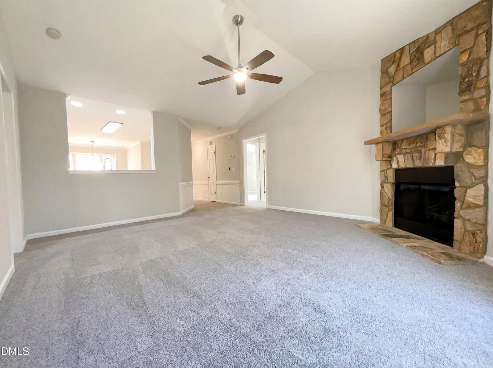 A spacious living room with light gray carpet, white walls, a ceiling fan with lights, and a stone fireplace. There is a cut-out window to the kitchen and doorways leading to other rooms. Natural light fills the space.