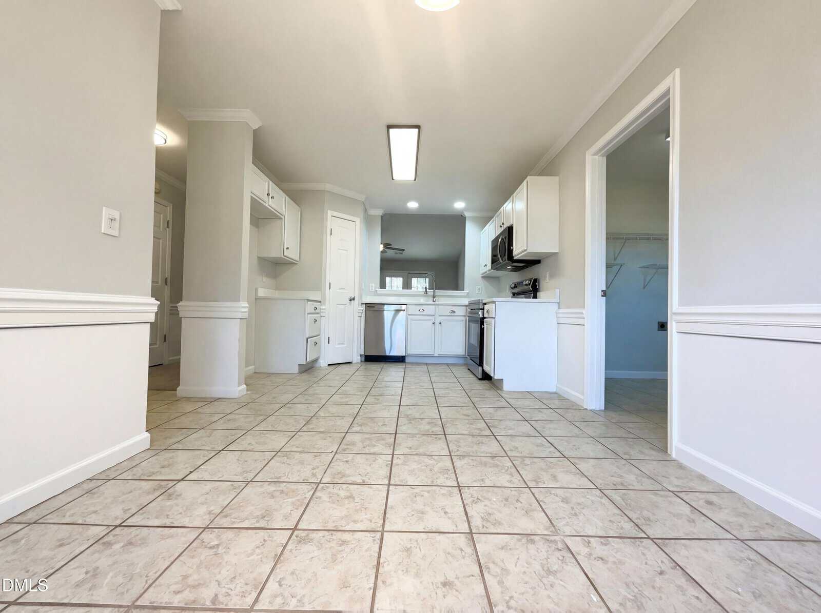 A spacious kitchen with white cabinets, stainless steel appliances, tiled floor, and neutral-colored walls. A doorway on the right leads to a laundry area with shelves, and there is recessed lighting on the ceiling.