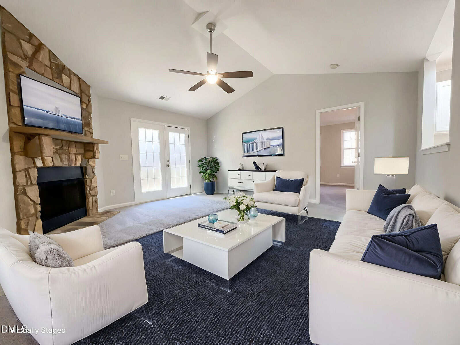 A bright living room with white sofas, a dark blue rug, a coffee table with flowers, a stone fireplace with a TV above it, a ceiling fan, and French doors leading outside. Walls are light gray with white trim.