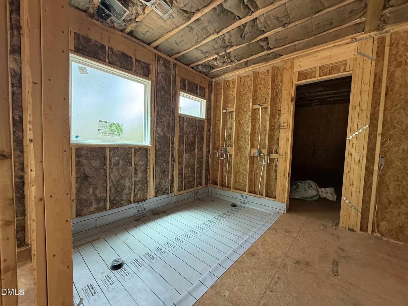 Bathroom under construction with exposed wooden framing, insulation in the walls and ceiling, two windows, plumbing fixtures, and a large shower base installed on the floor. No drywall or finished surfaces yet.