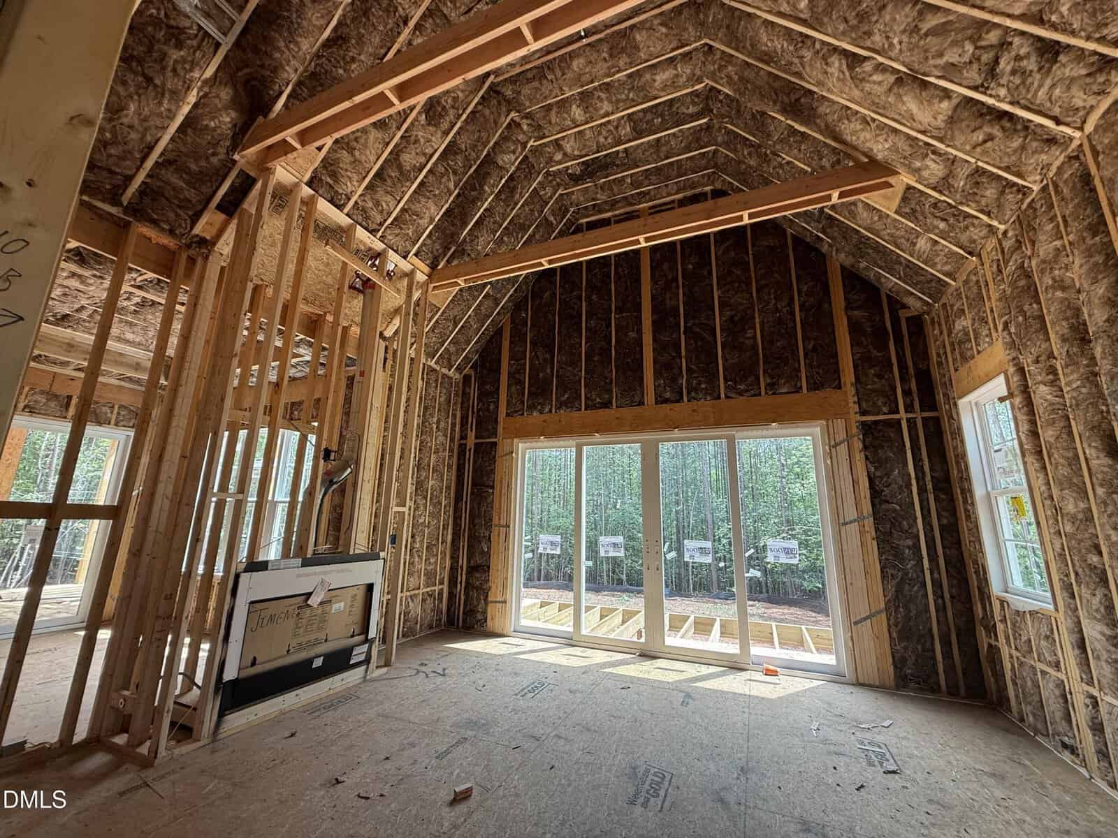 A room under construction with exposed wooden framing and spray foam insulation, large windows and glass doors facing a wooded area, and unfinished floors and ceiling.