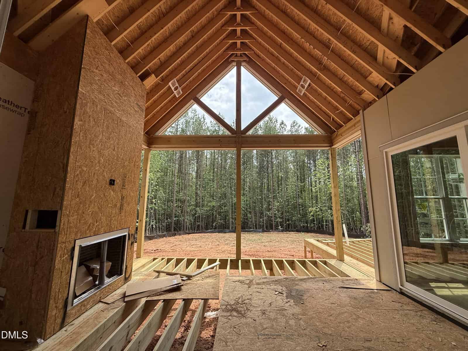 View from inside a house under construction, showing exposed wooden beams and large windows overlooking a forested area with trees and cleared red dirt ground outside.