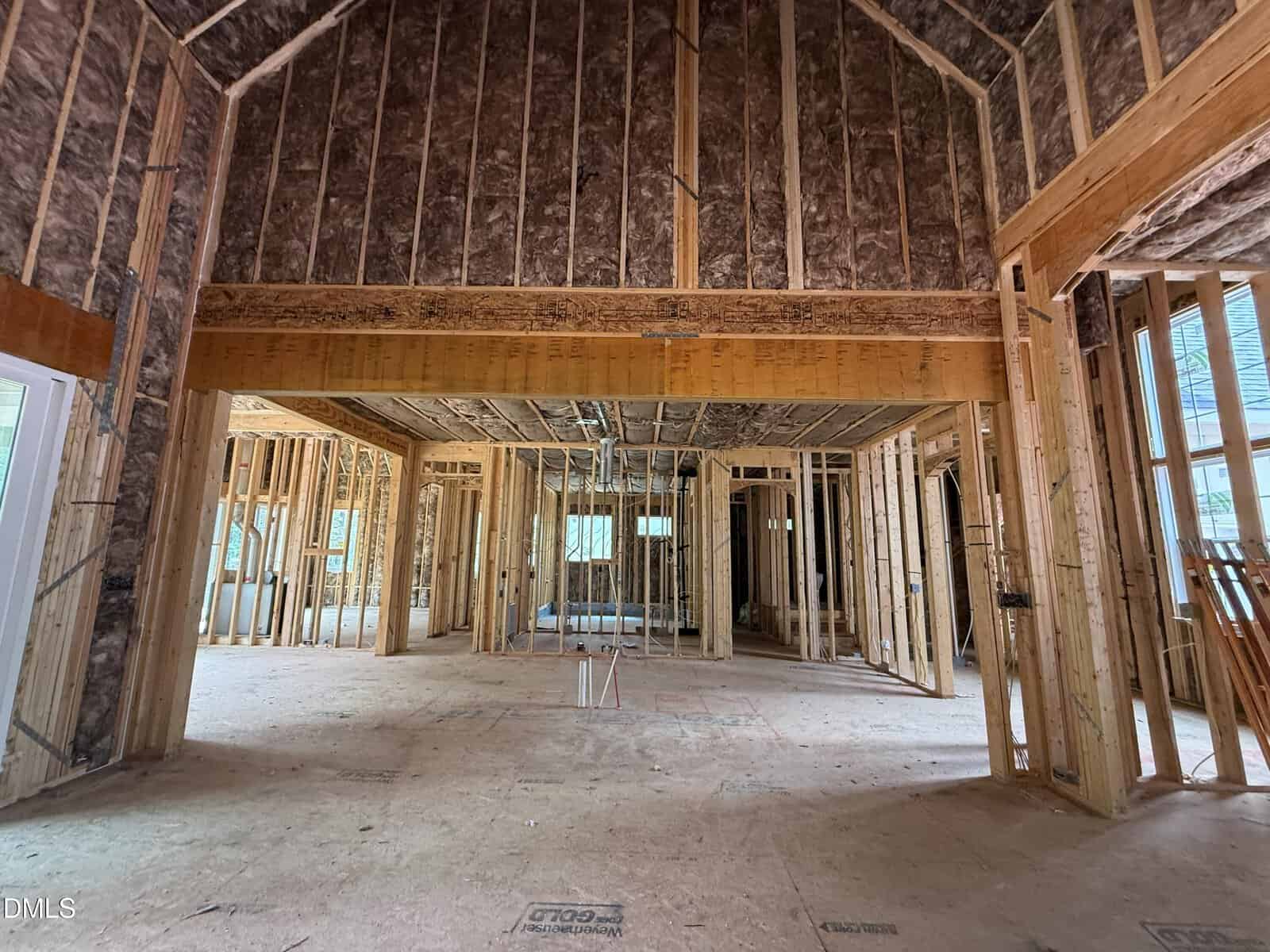 Interior view of a house under construction, with exposed wooden framing, insulation in the ceiling, and unfinished floors and walls. No drywall or electrical fixtures are installed yet. Natural light enters from windows.