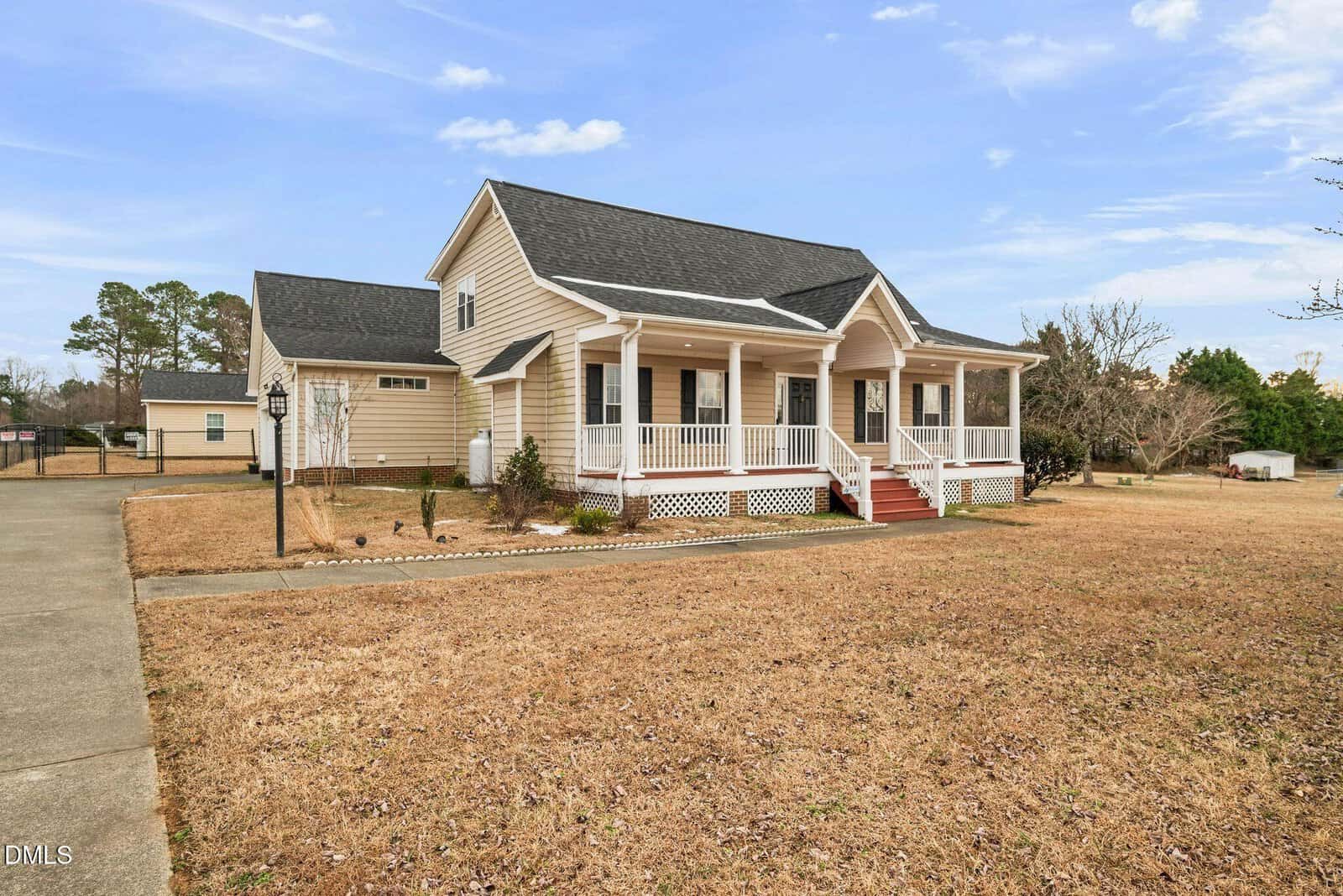 A beige, single-family house with a front porch and white railings sits on a large, dry lawn. The house has a black roof and red steps leading to the porch. A concrete driveway is visible on the left side.