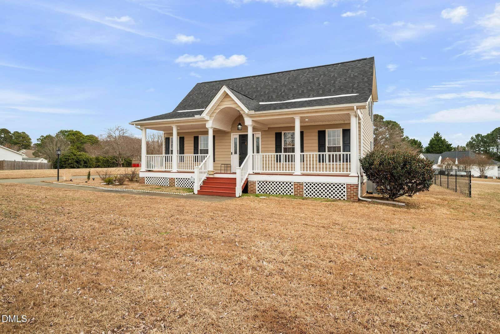 A single-story beige house with a black roof, red steps, and a covered front porch sits on a large, dry lawn. White railings border the porch, and there are a few bushes and trees in the background under a blue sky.