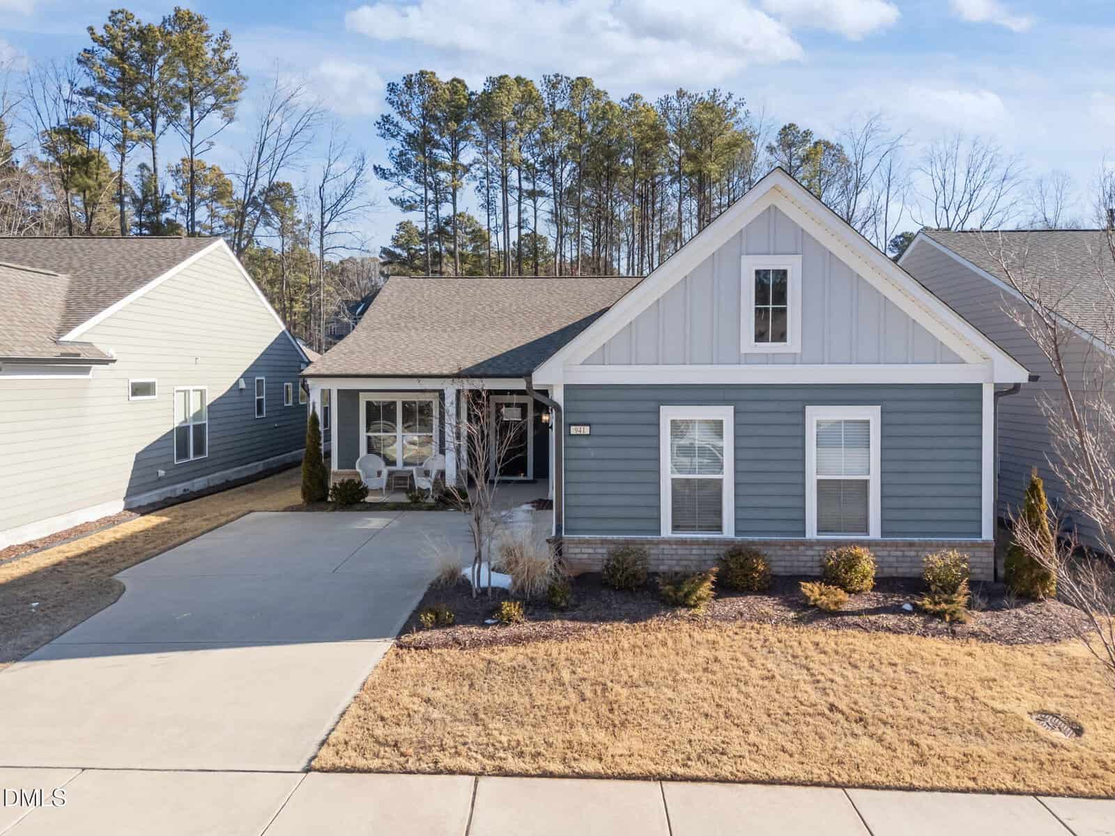 A single-story blue house with white trim, front porch, and gable roof sits between two similar homes. A driveway leads to the entrance. Brown grass and minimal landscaping are visible in the front yard. Tall trees are in the background.