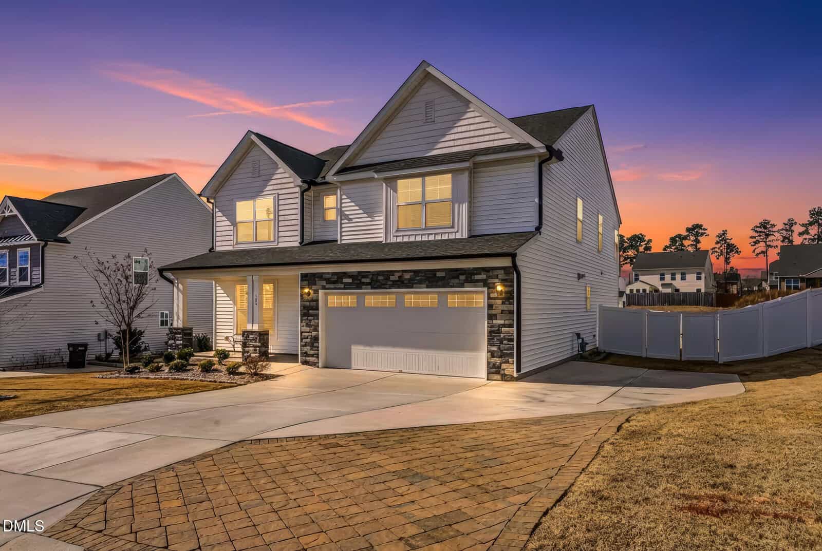 Two-story suburban house with white siding, a covered front porch, and a two-car garage. Driveway and walkway are visible, with exterior lights on at sunset. Neighboring homes and trees appear in the background.