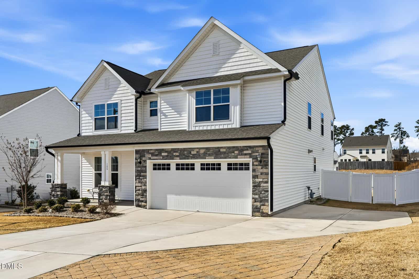 Two-story suburban house with white siding, dark roof, and a double garage. The driveway is curved and paved. There is a fenced backyard and a small lawn with minimal landscaping. The sky is clear and blue.