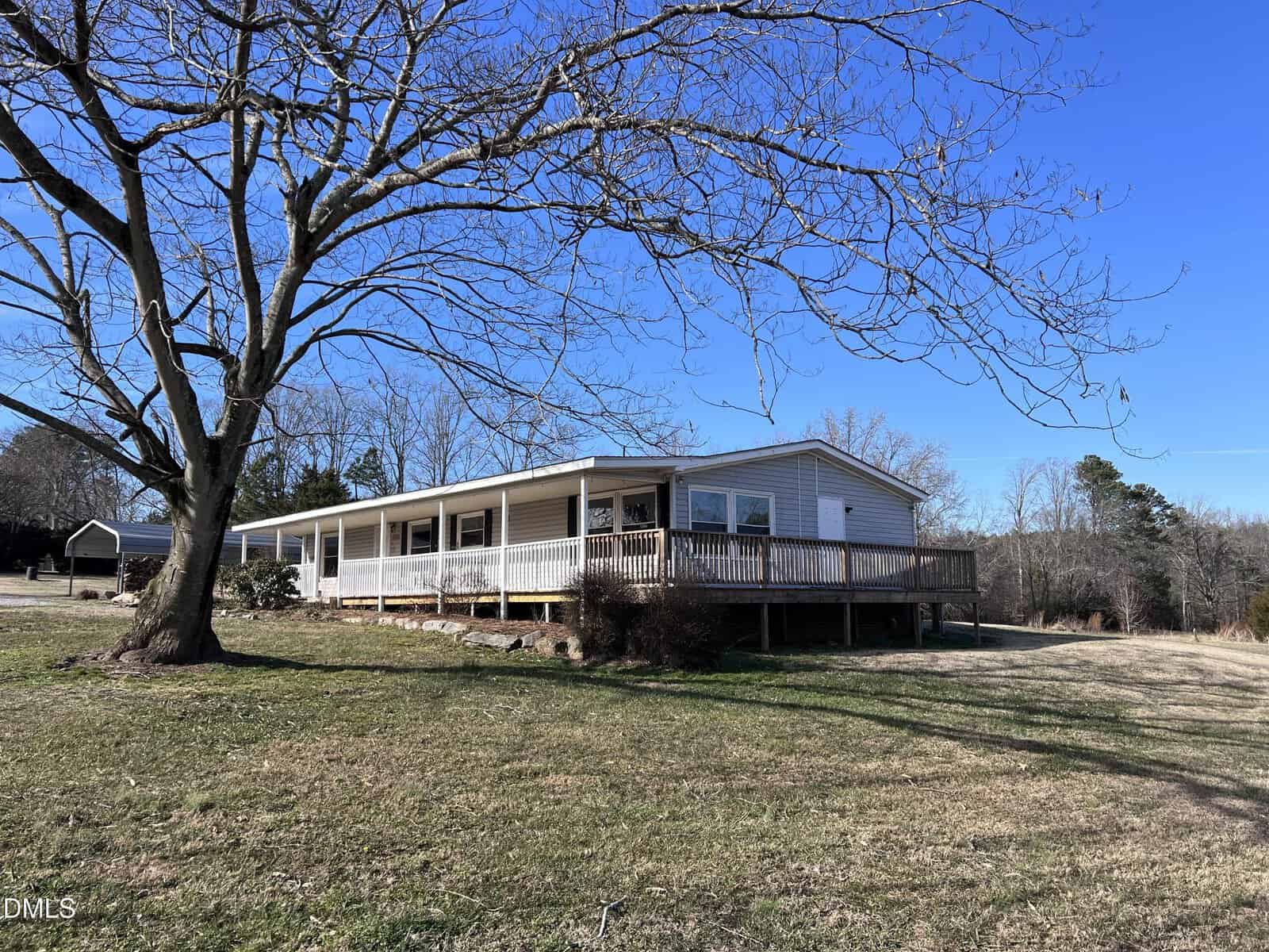 A single-story house with a wide front porch and wooden ramp sits on a grassy lot. A leafless tree is in the foreground, and the sky is clear and blue. Trees and a neighboring house are visible in the background.