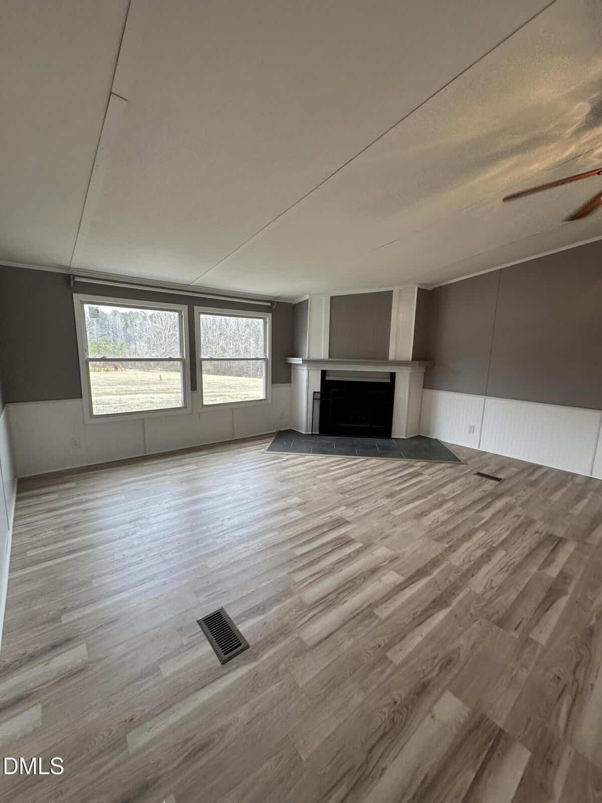 A spacious, empty living room with light wood flooring, gray and white walls, large windows with a view of trees, and a corner fireplace with a white mantle. A ceiling fan is partially visible.
