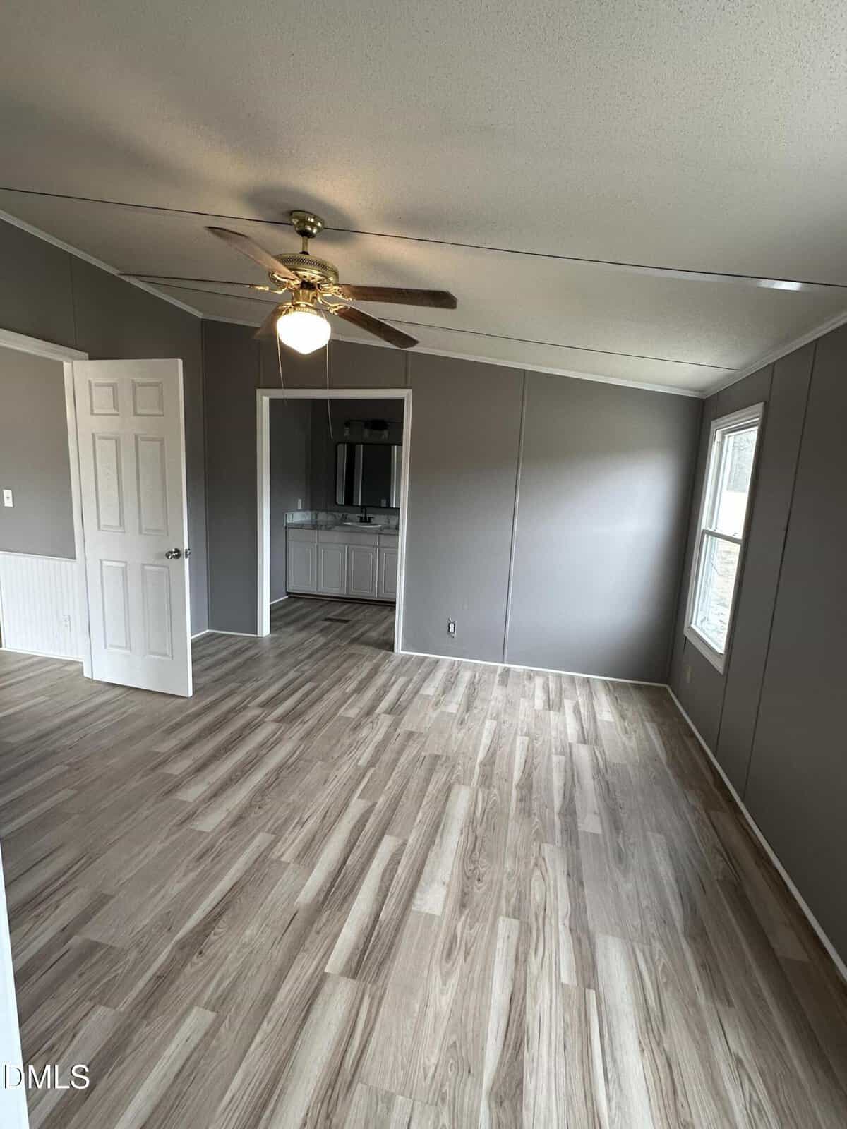 A mostly empty room with light wood flooring, gray walls, a ceiling fan with a light, an open door leading to a bathroom with a double sink, and a window letting in natural light.