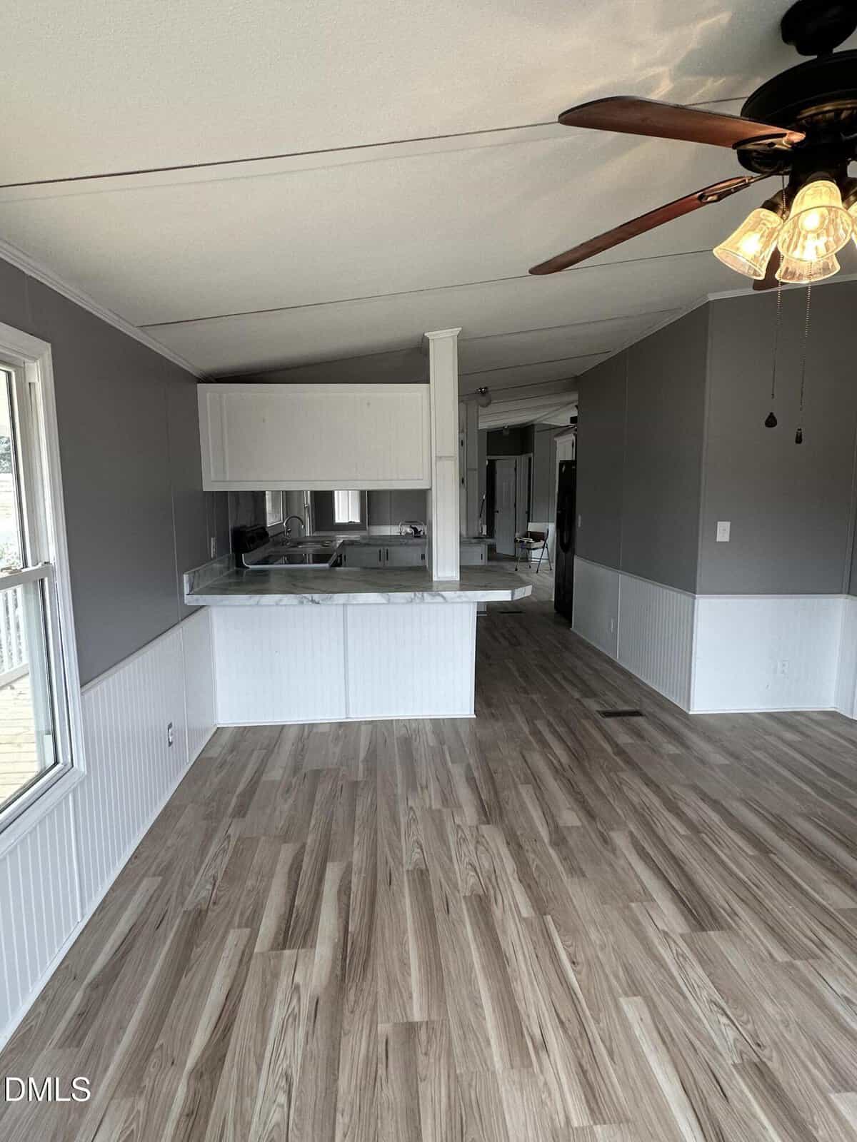 Room with wood-style flooring, gray and white walls, ceiling fan with lights, large window, and a kitchen area with marble countertop and white cabinets visible in the background.