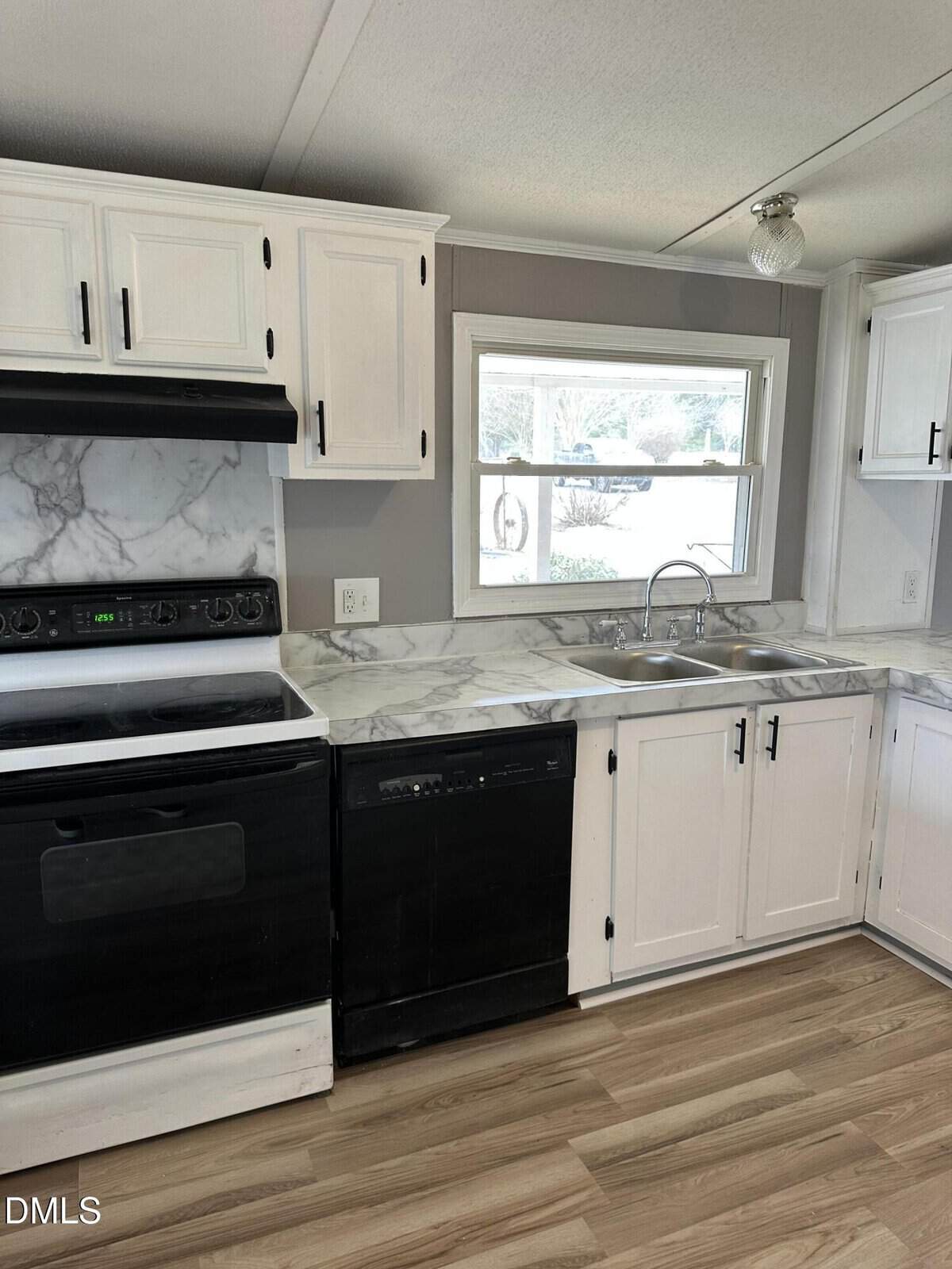 A kitchen with white cabinets, marble-pattern countertops, a black stove with a matching range hood, a black dishwasher, a double sink under a window, and light wood flooring.