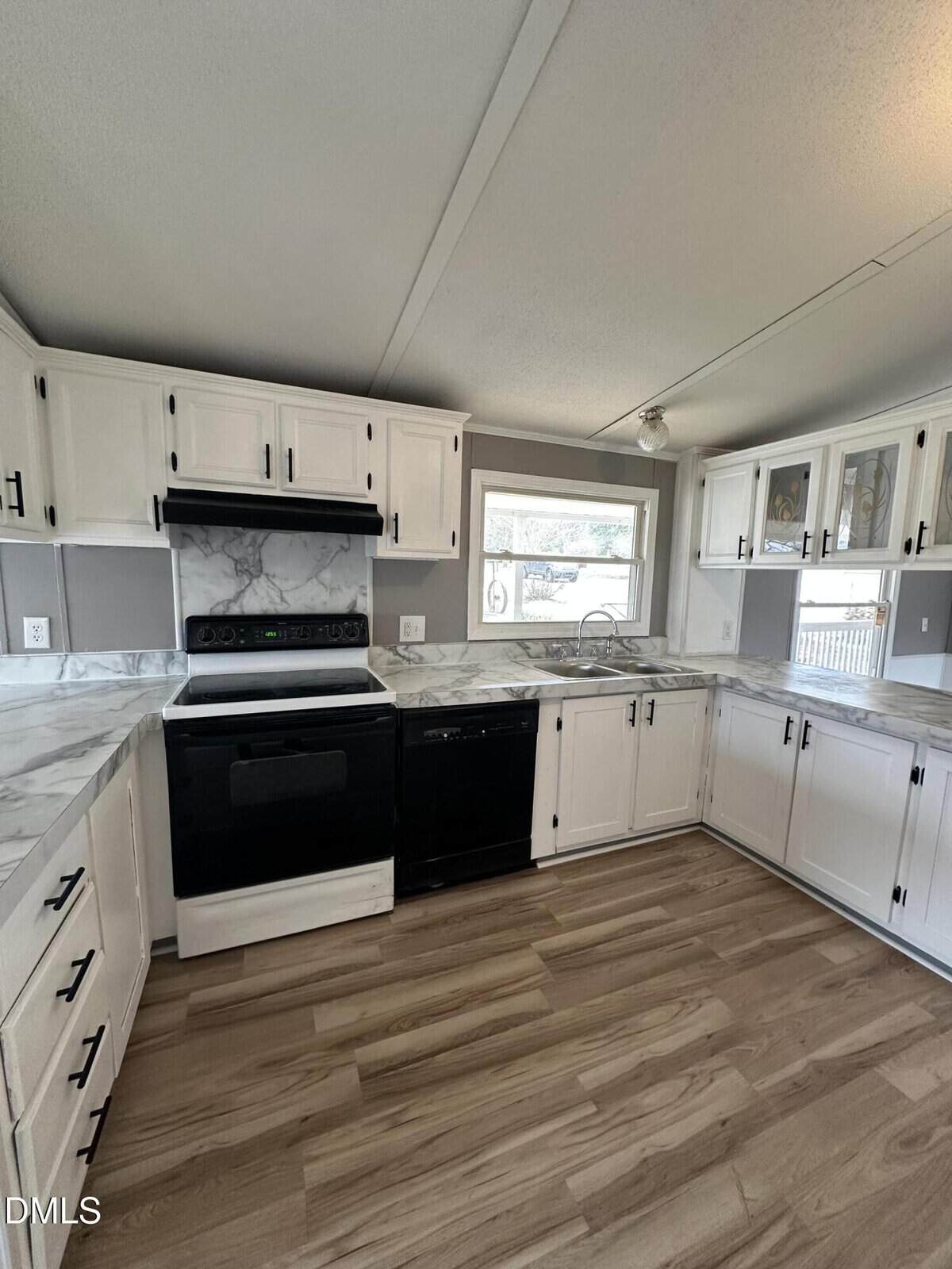 Kitchen with white cabinetry, marble-look countertops, black stove, black dishwasher, and wood-look flooring. There is a window above the sink and upper cabinets with glass inserts on the right.