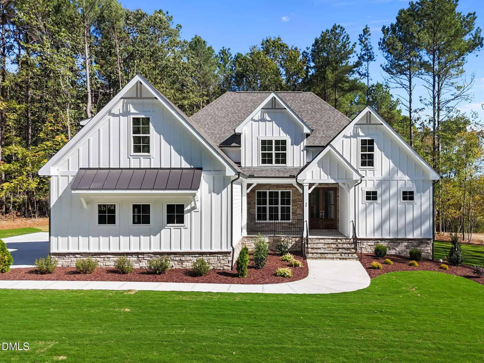 A two-story white house with black trim, stone accents, and a covered front porch. The home is surrounded by a green lawn, landscaped bushes, and trees in the background under a clear blue sky.