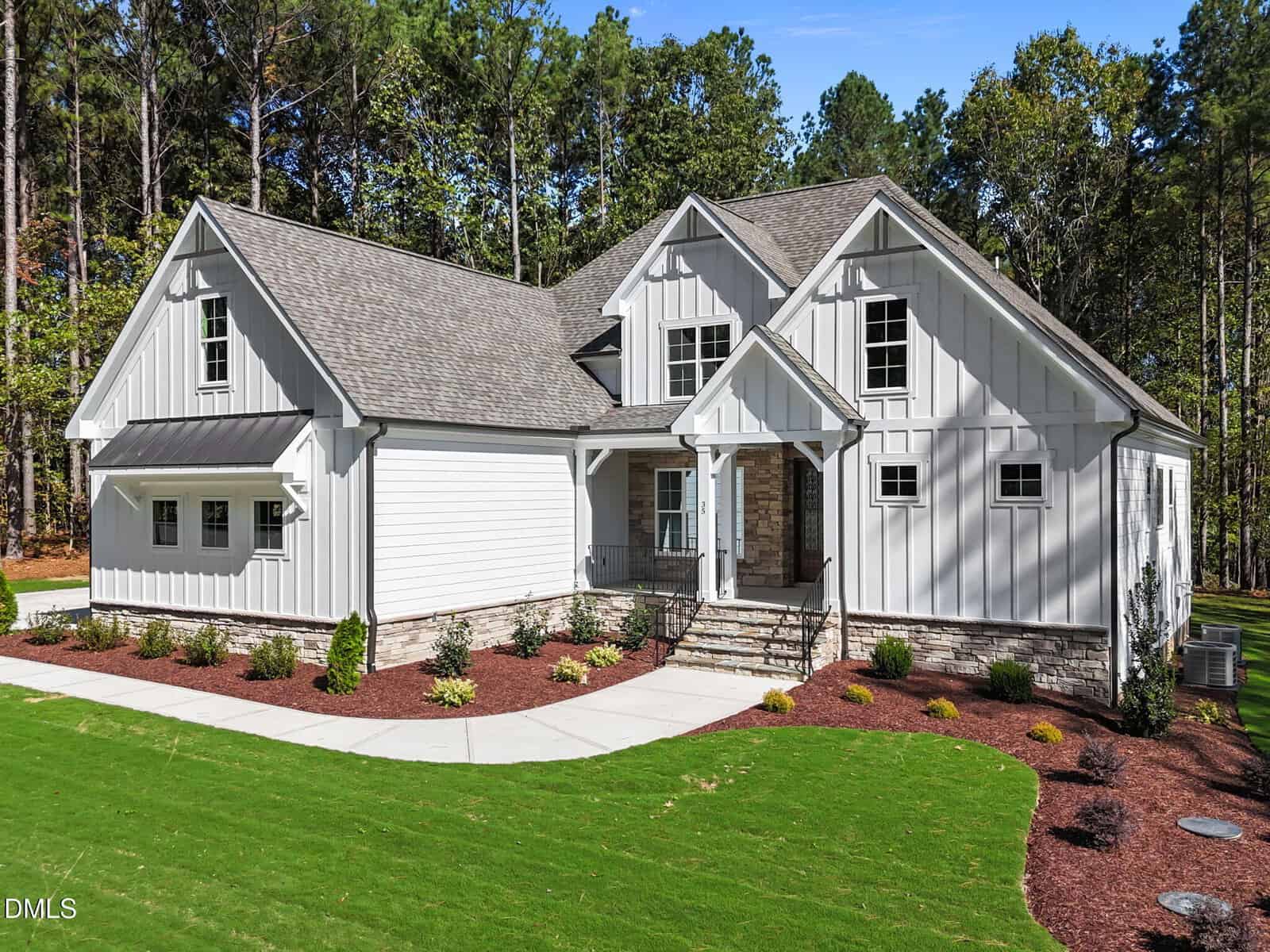 A two-story modern farmhouse with gray siding, gabled roof, stone accents, covered entryway, and landscaped yard, set against a backdrop of tall trees and a well-maintained lawn.