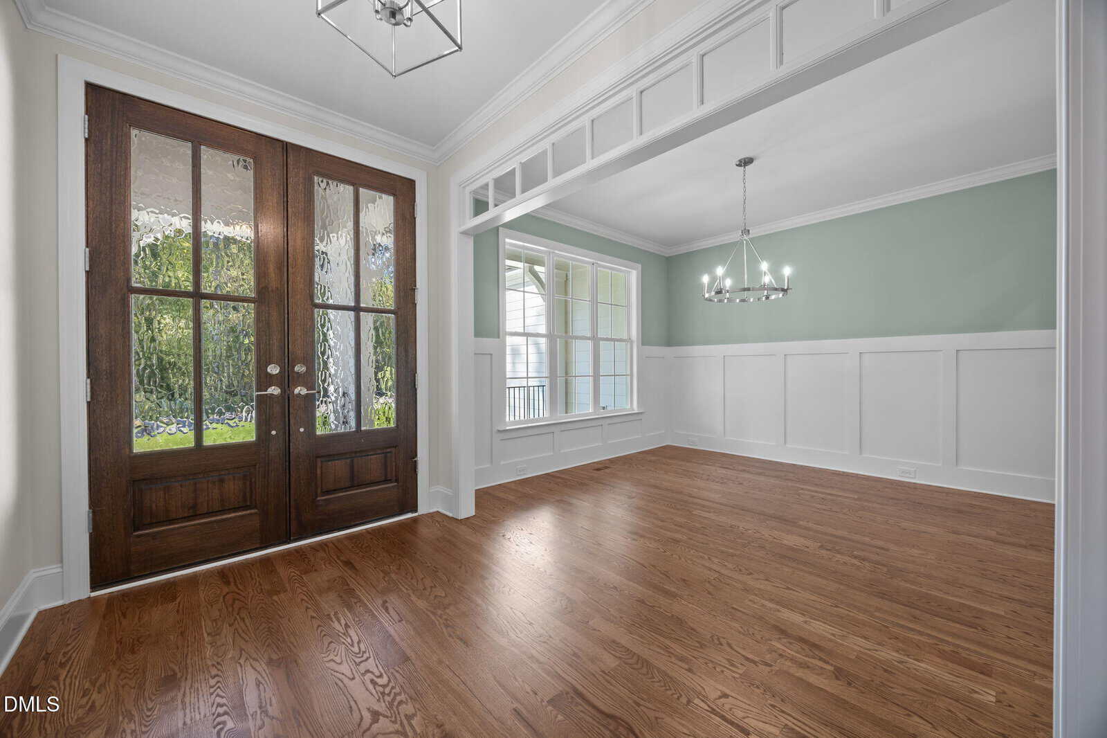 Spacious empty room with light wood flooring, pale green walls with white wainscoting, large windows, a double wooden door, and a modern chandelier hanging from the ceiling.