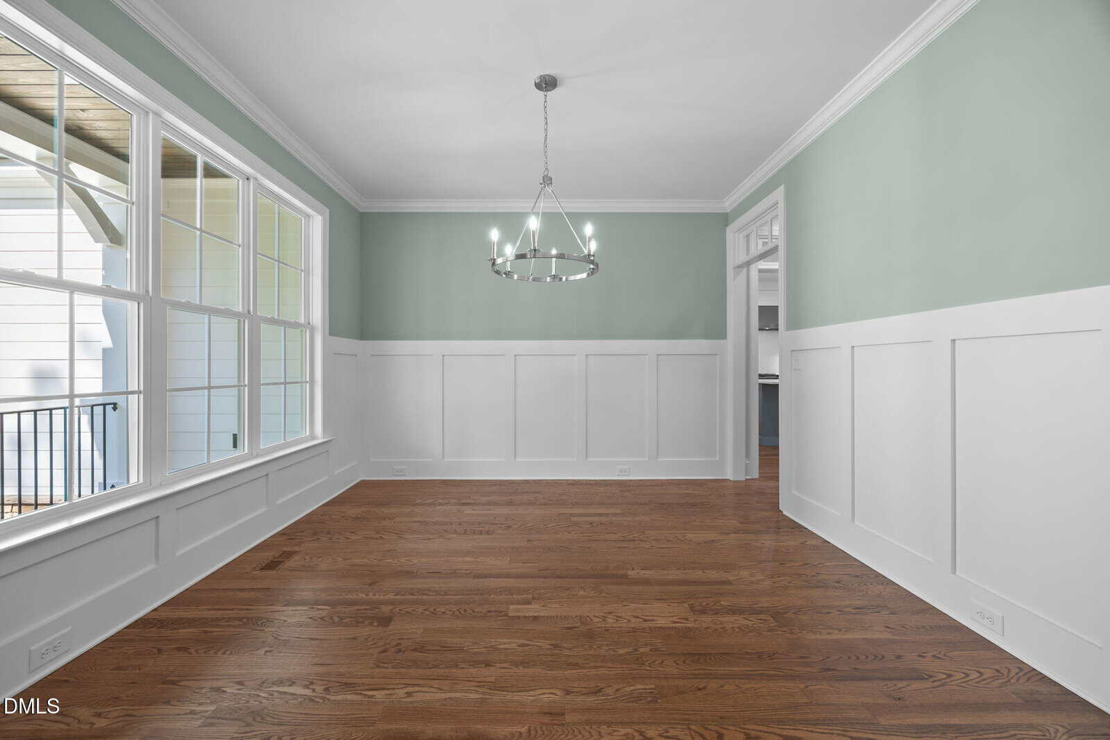 Empty dining room with light green upper walls, white wainscoting, a modern circular chandelier, large windows on the left, and hardwood floors. An open doorway leads to an adjacent room.