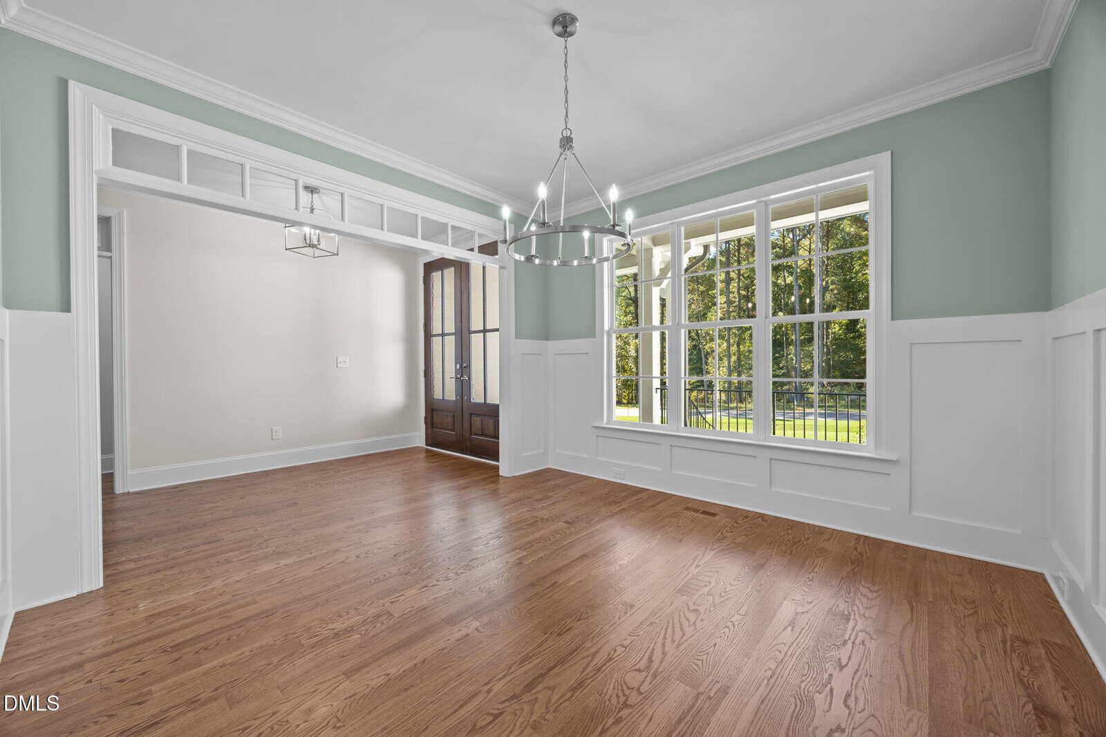 Bright, empty dining room with wood floors, white wainscoting, green walls, large windows showing trees and grass outside, and a modern chandelier hanging from the ceiling.