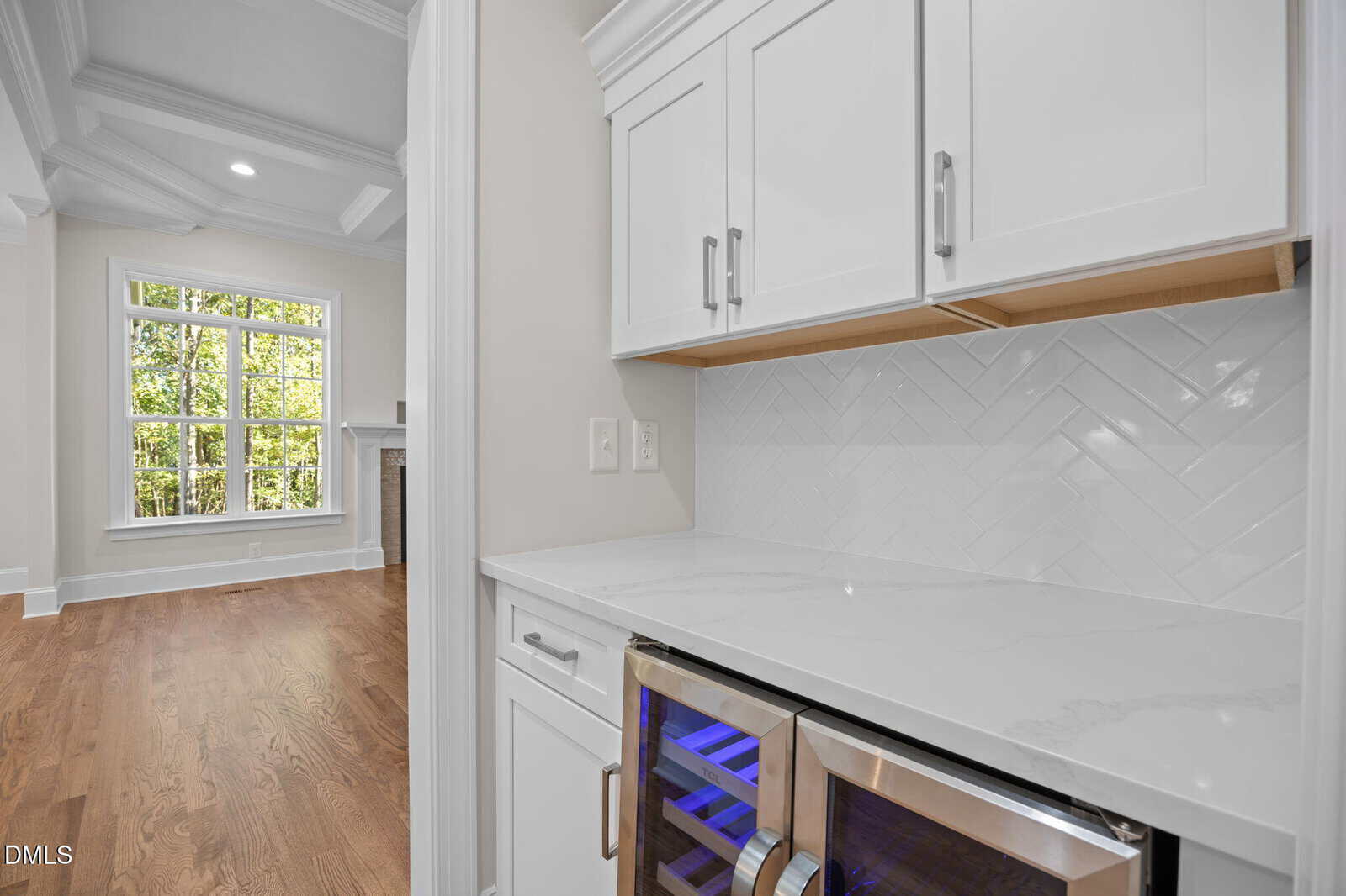 A kitchen nook with white cabinets, a marble countertop, herringbone tile backsplash, and a double glass-door wine fridge. Hardwood floors run throughout, and a large window is visible in the adjacent room.