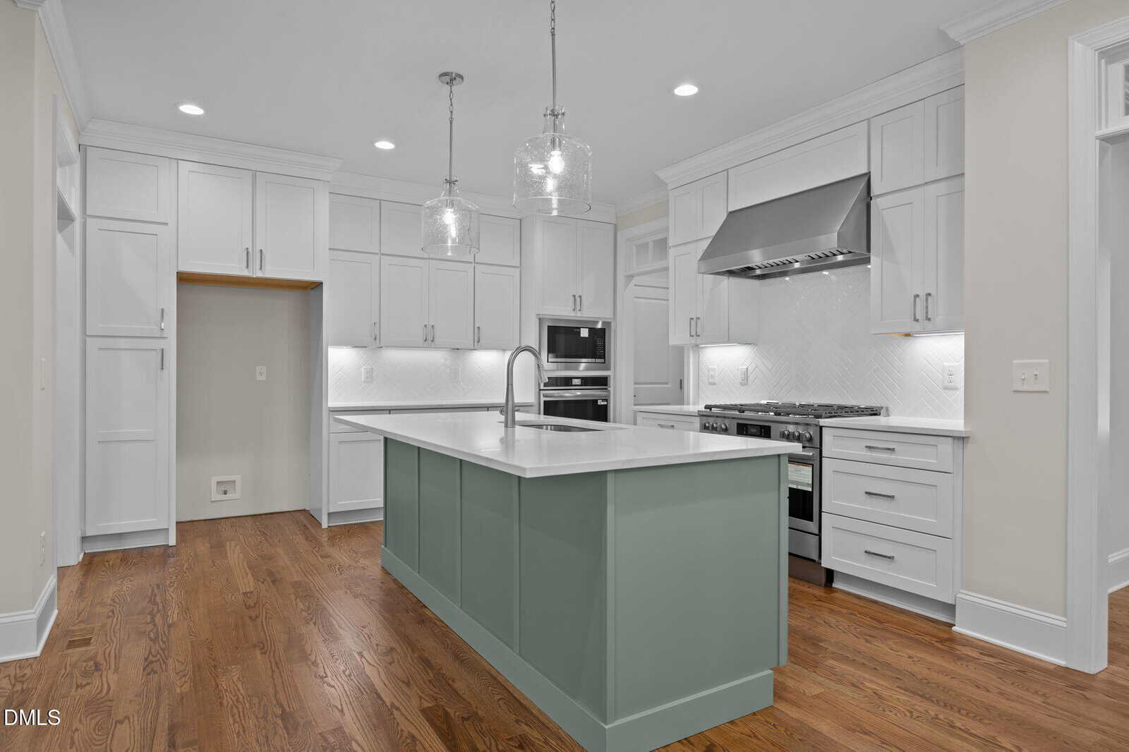 Modern kitchen with white cabinets, stainless steel appliances, a green island with a sink, pendant lights overhead, hardwood floors, and white tile backsplash. Walls are light beige, and recessed lighting is present.