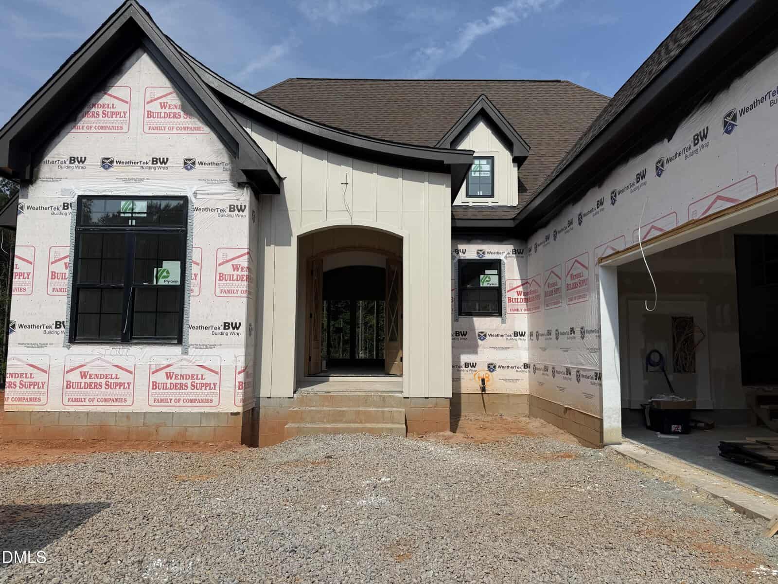 A house under construction with black window frames, unfinished walls, exposed insulation, and an open garage on the right, set on a gravel and dirt driveway.