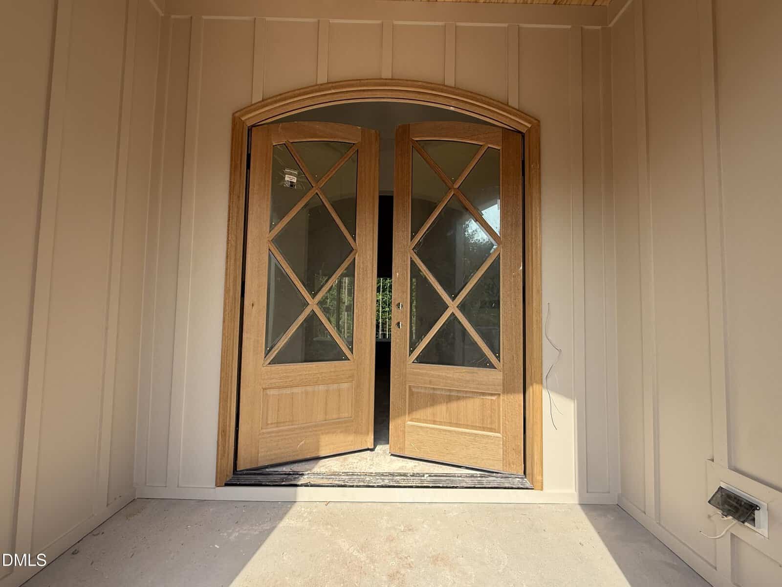Wooden double doors with glass panels are open, revealing an entryway with sunlight streaming in. The doors have an arched top and a crisscross design on the glass. The walls are beige with vertical panels.