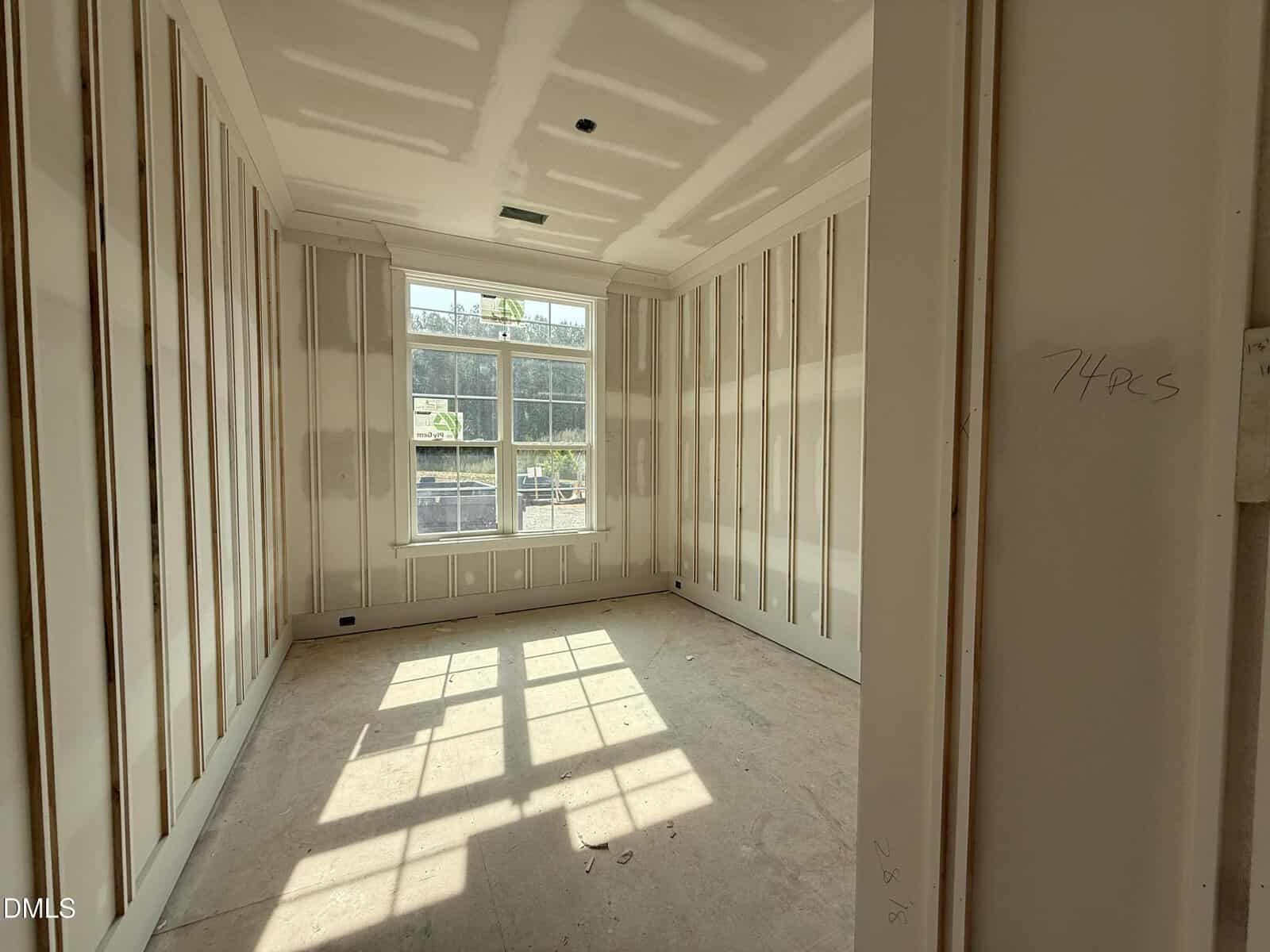 Sunlit, unfinished room with drywall and exposed seams, large window letting in bright light, bare floor, and construction notes written on the wall.