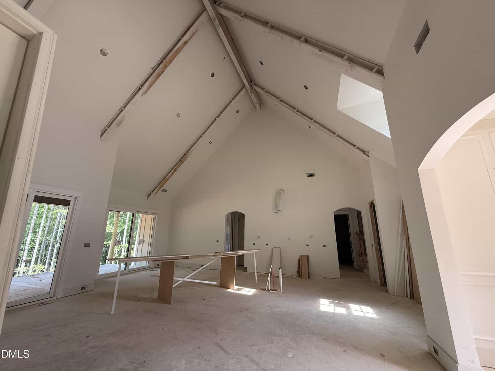 Spacious room under construction with vaulted ceiling, exposed beams, unfinished floors, large windows, and scattered building materials. Sunlight streams through the windows, illuminating the interior.