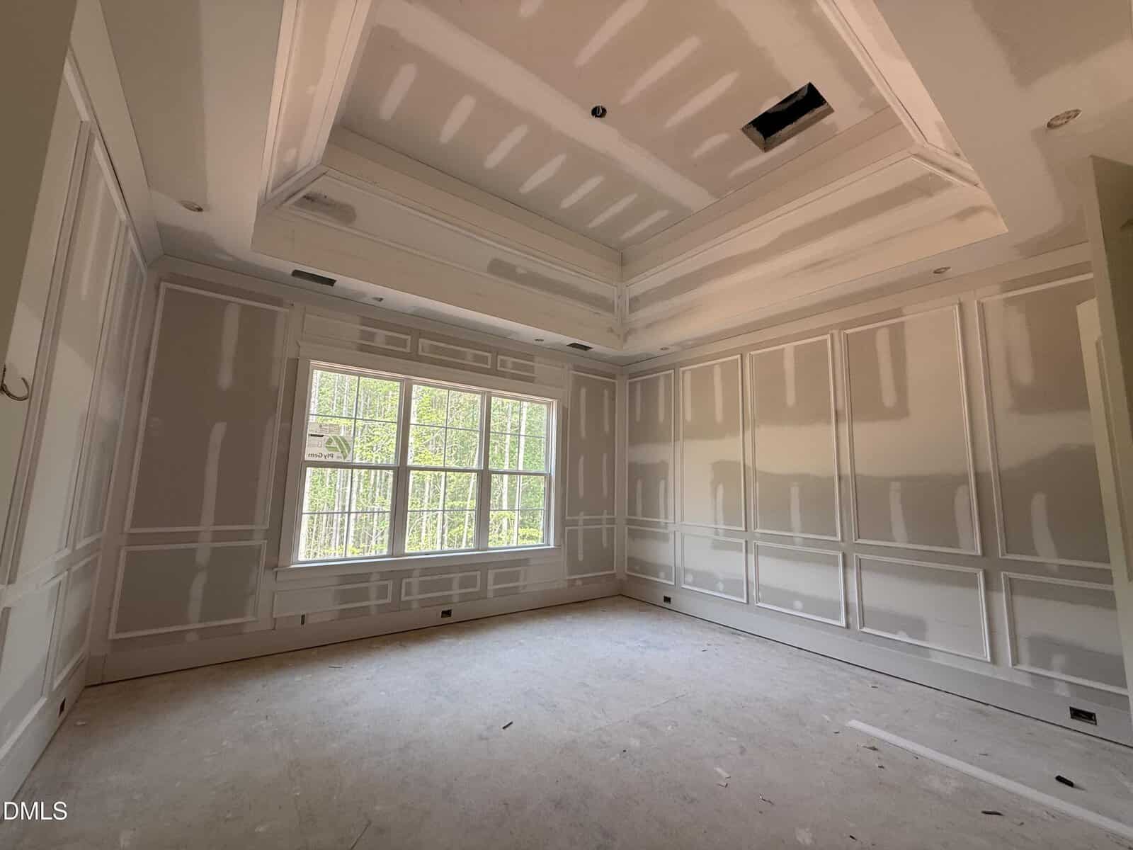 An empty room with unfinished drywall, large windows letting in natural light, and a ceiling with exposed seams. The floor is bare, and no fixtures or furniture are present.