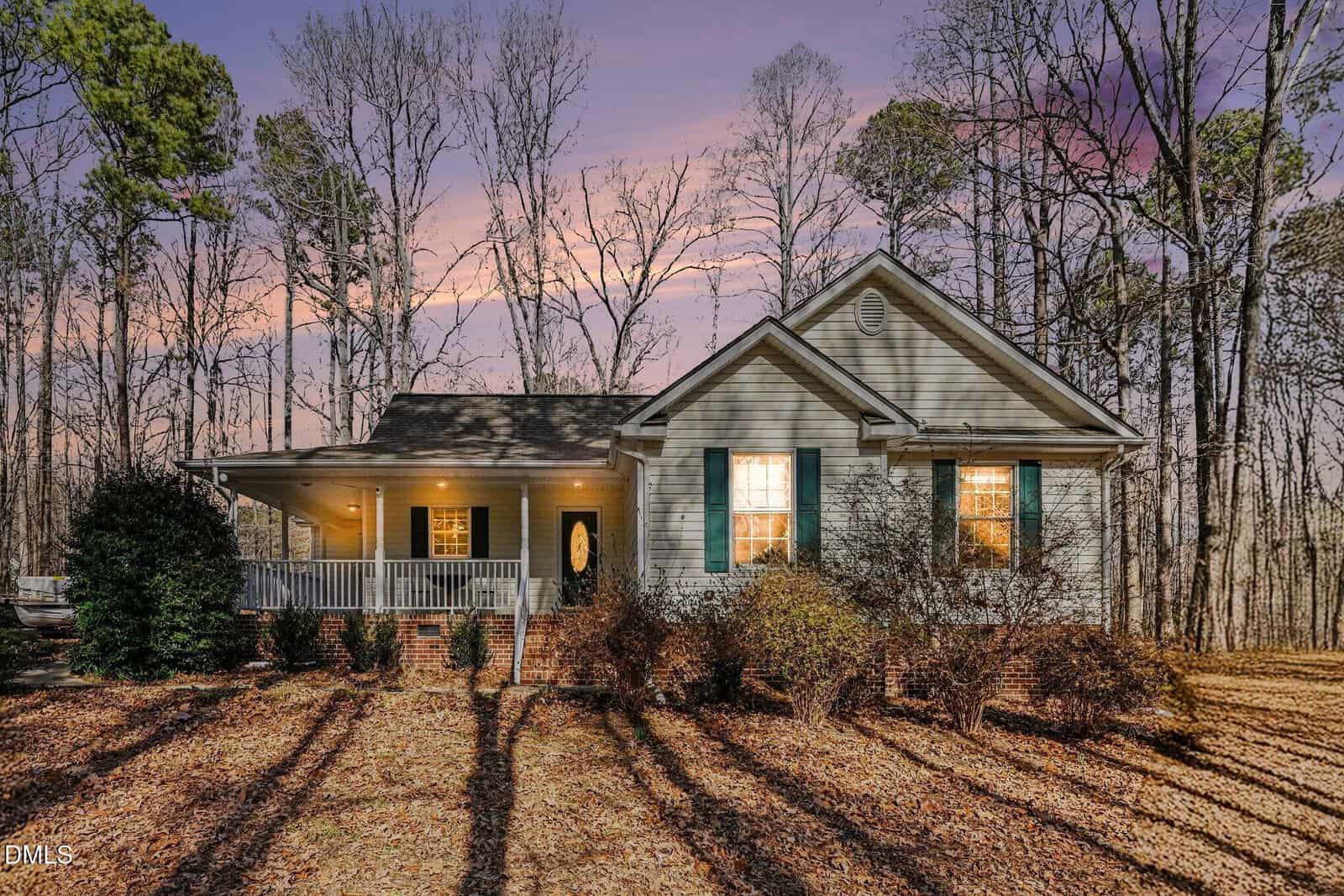 A single-story house with a large front porch, green shutters, and a glass front door is surrounded by leafless trees at sunset. Warm lights illuminate the porch and interior, with fallen leaves covering the yard.