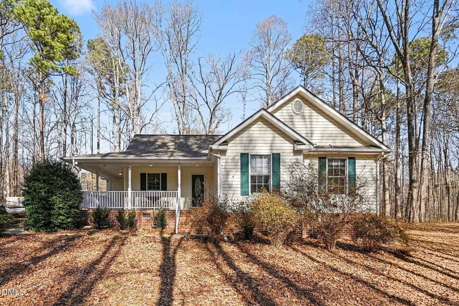 A single-story house with beige siding, green shutters, and a front porch with white railings, surrounded by tall trees and leaf-covered ground under a clear sky.